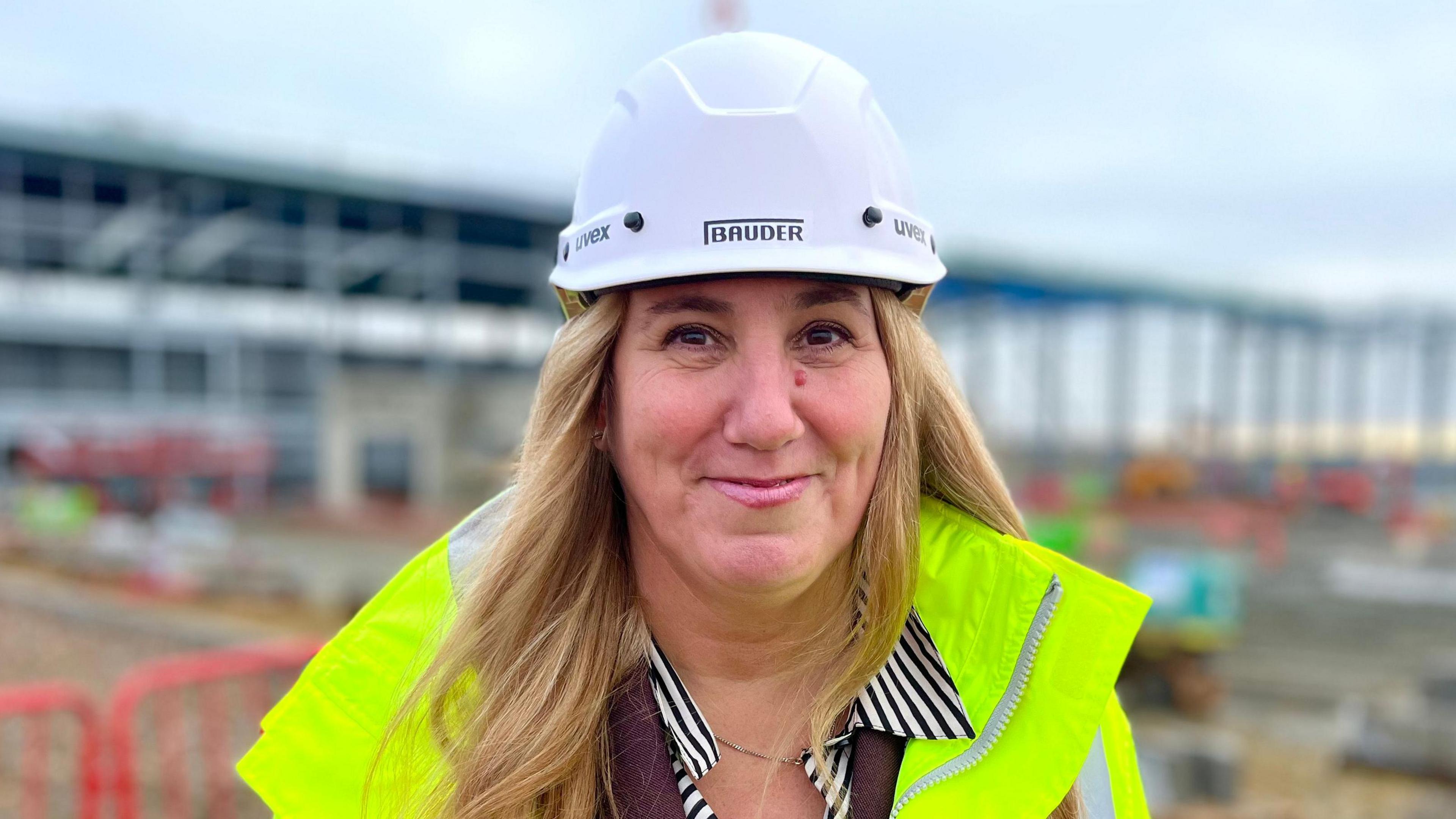 Yvonne Higgins smiles at the camera. She is wearing high-vis clothing and a hard hat which has "Bauder" written on it. She is on a building site, which can be seen behind her. 