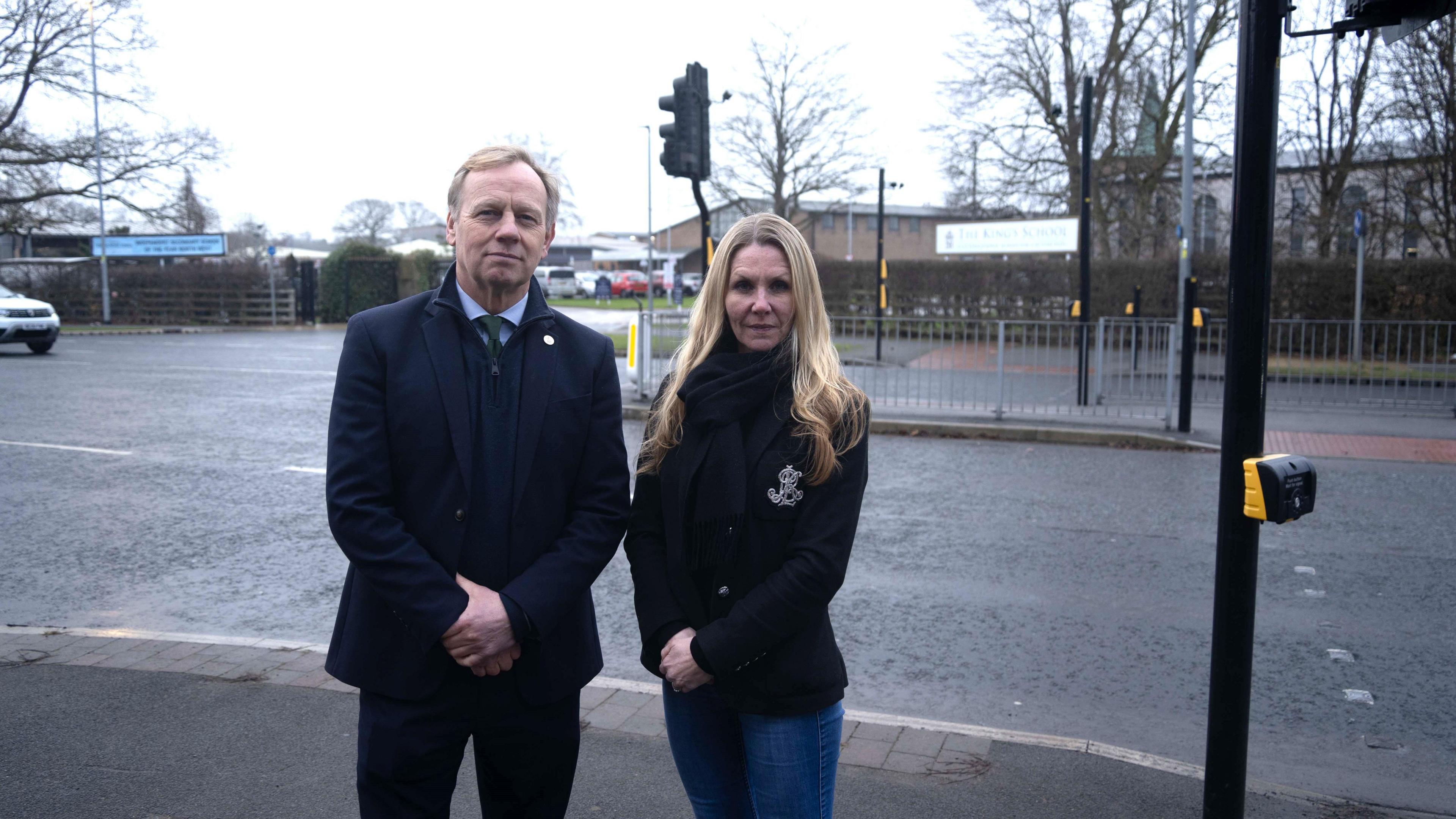 George Hartley and Claire Taylor stand at the side of the road by the crossing outside the school. Mr Hartley is wearing a dark jacket, blue shirt and green tie and dark coloured trousers. Mrs Taylor wears a dark coloured blazer with jeans and a black scarf.. 