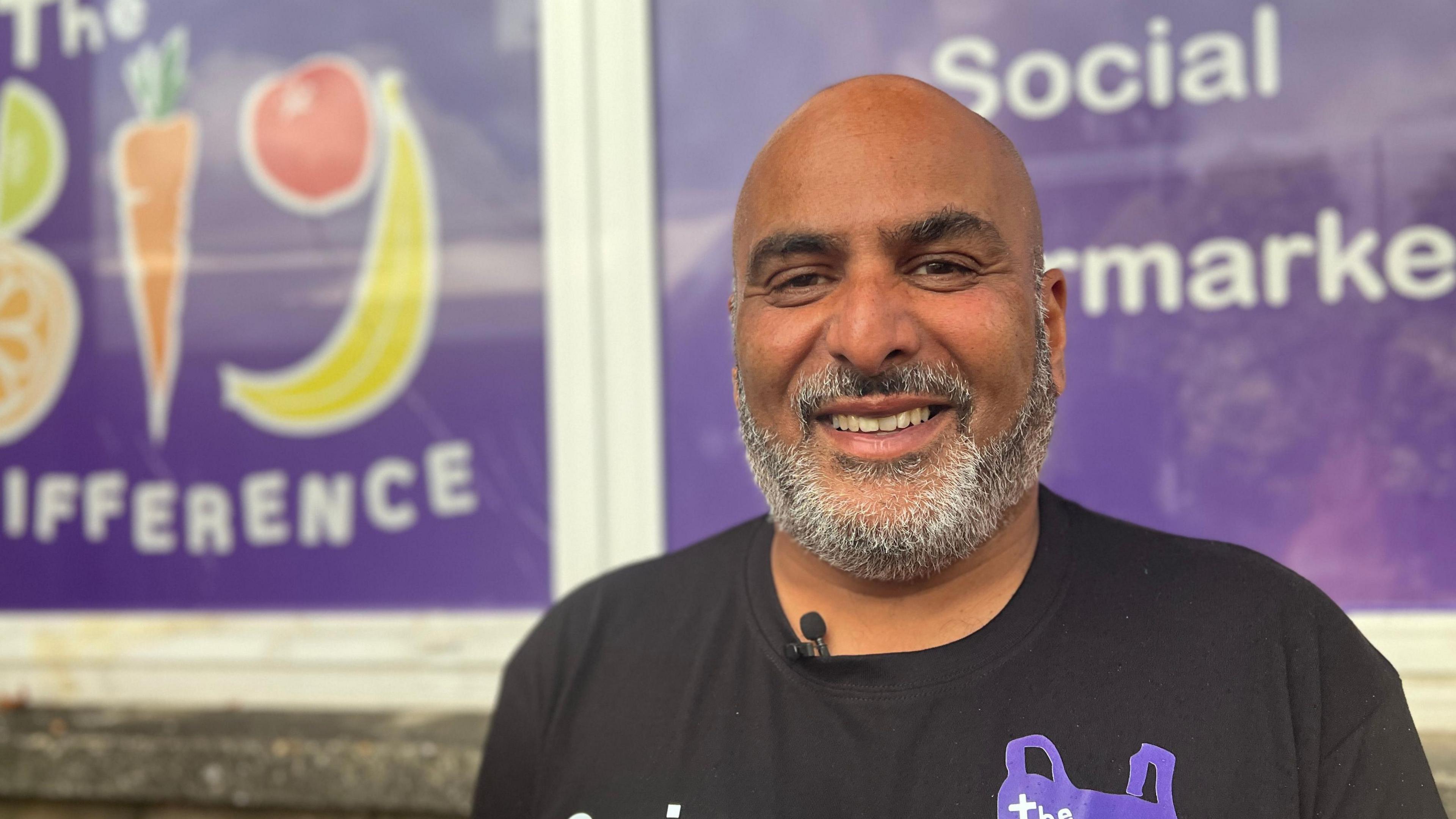Sanjay Mall looking into the camera. He is bald and has a gray beard and is wearing a black T-shirt with a purple shopping bag logo. In the background there is branding on the wall that says "The Big Difference" and "Social supermarket."