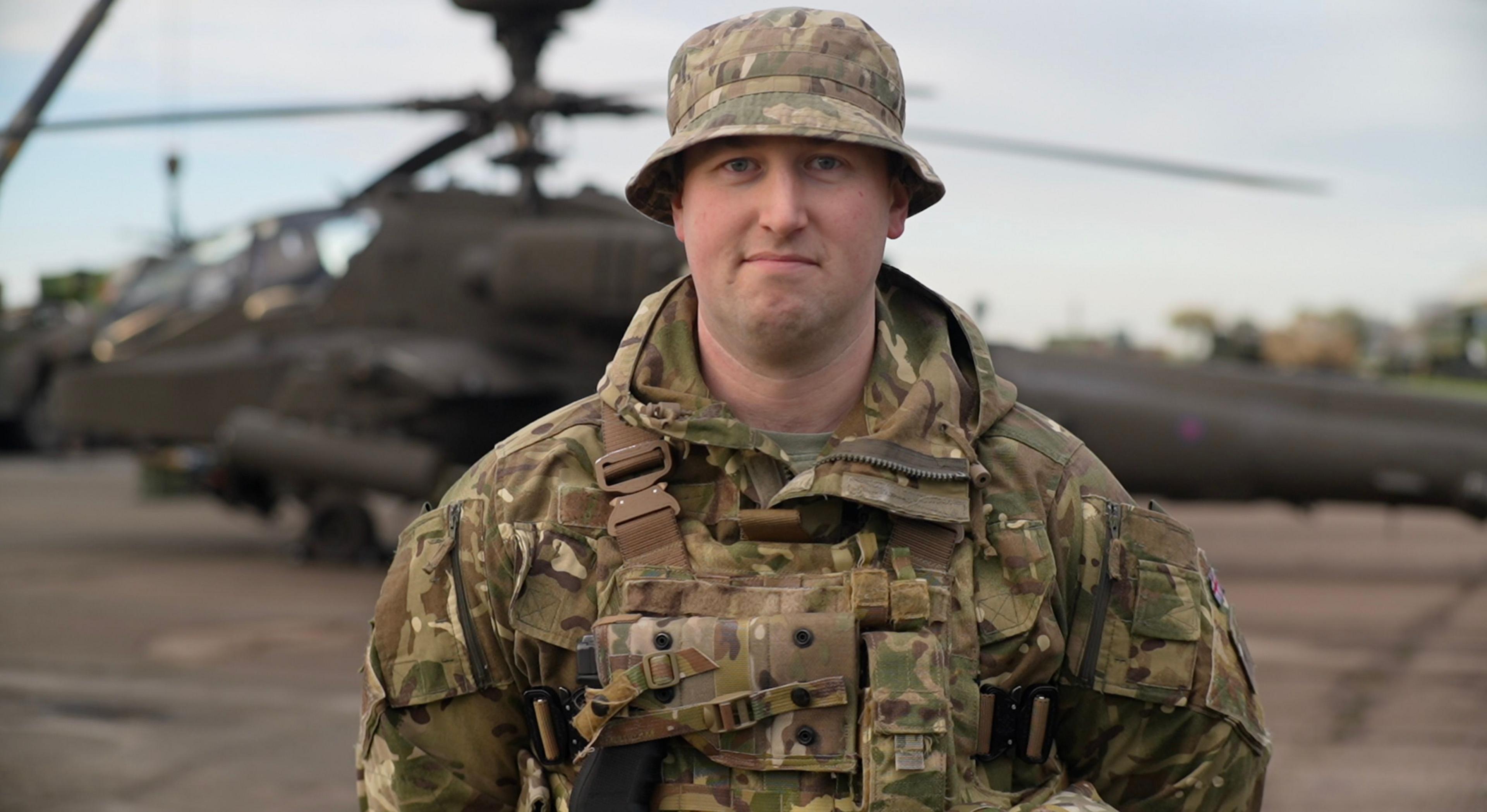 Mark smiles straight on at the camera. Helicopters rest on an airfield behind him. He wears a camouflage uniform along with a bucket hat.