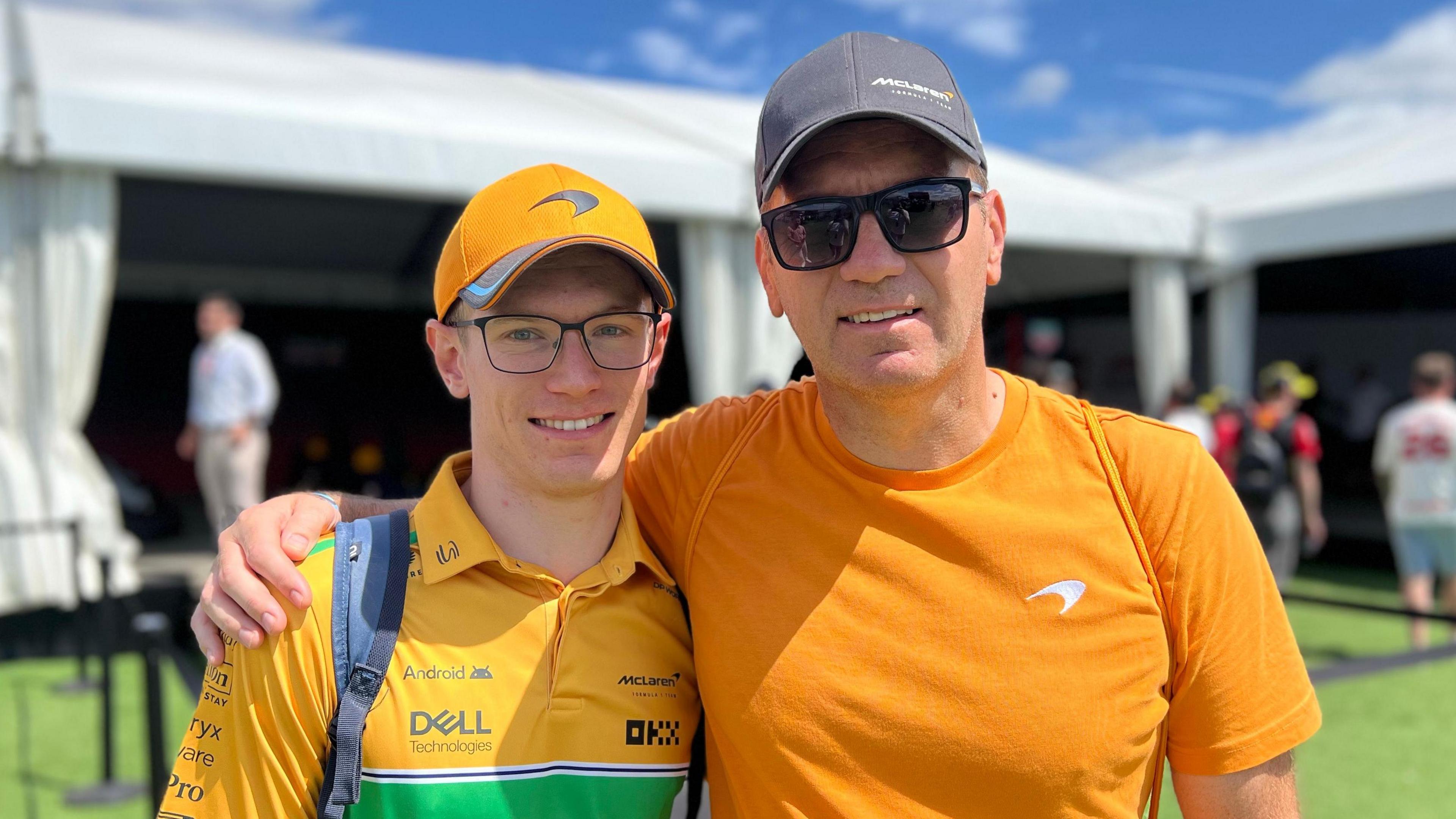 Two men in McLaren F1 apparel stand side-by-side at Silverstone Circuit on a sunny day.