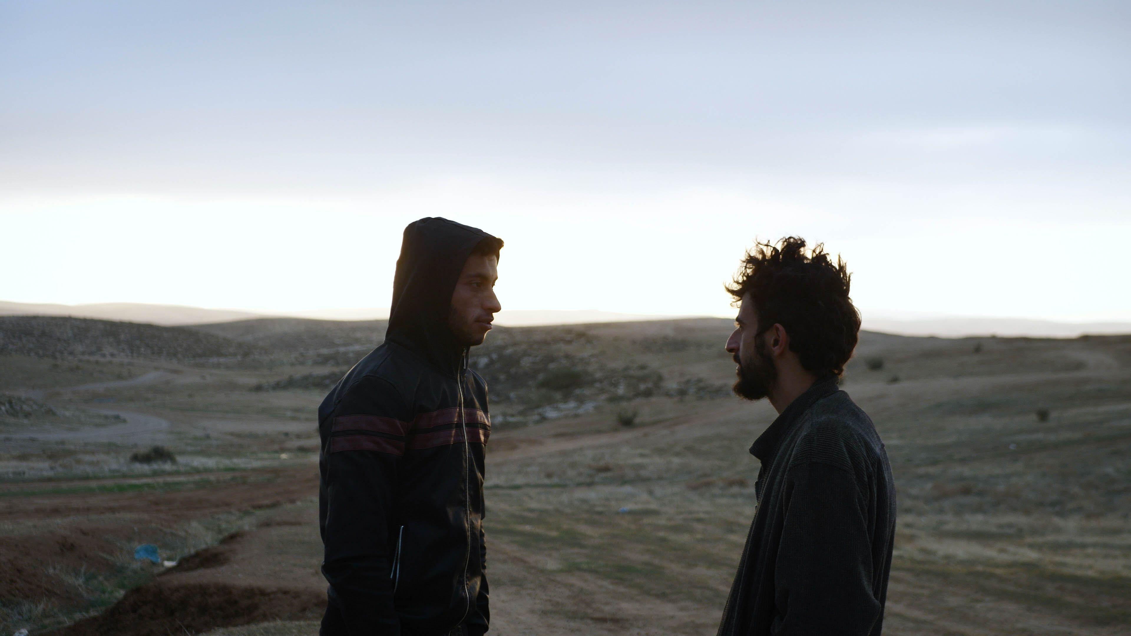 Two young men face each other and talk, one wearing a jumper with the hood up and the other a jacket, a barren, hilly terrain behind them