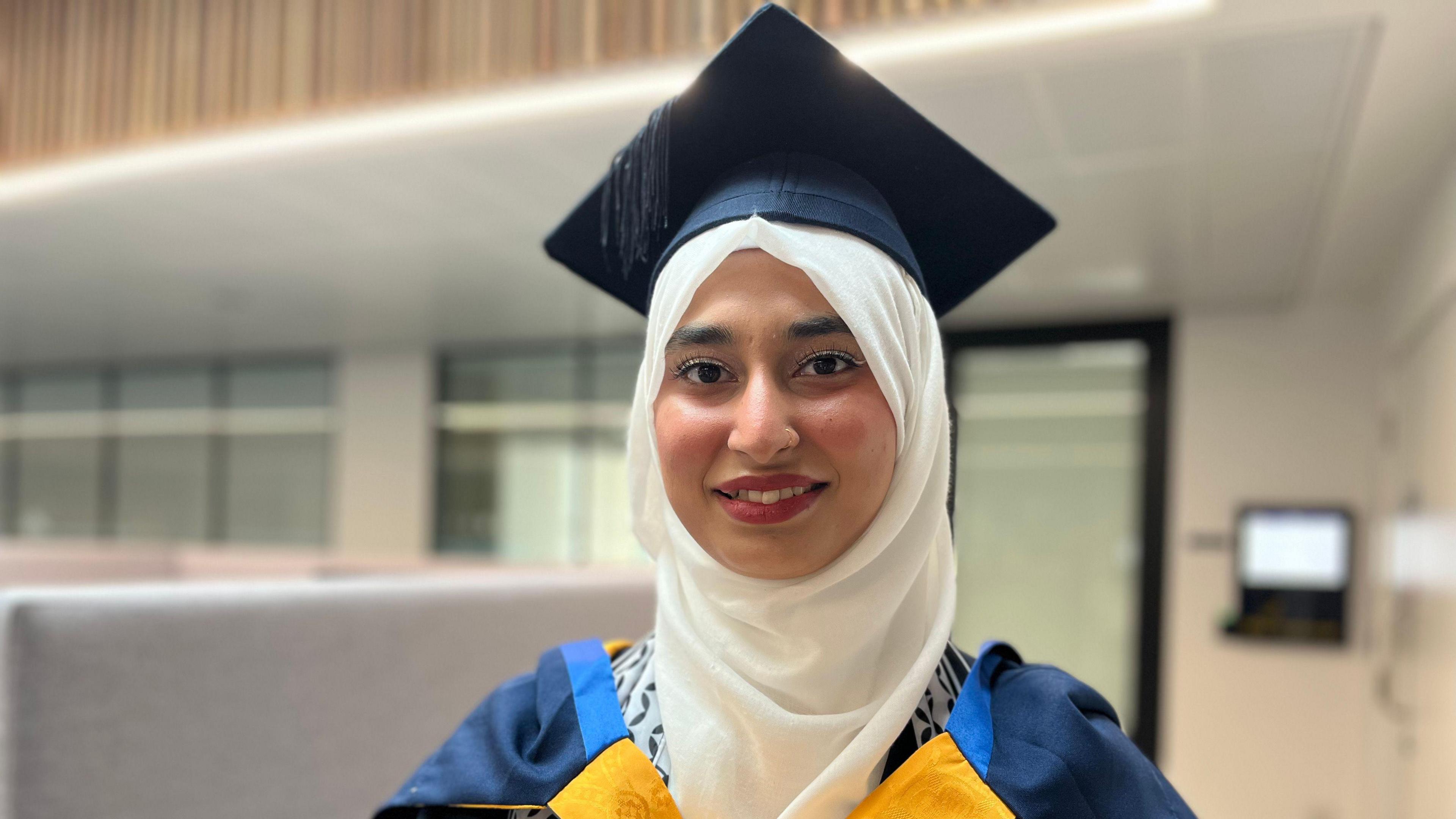 Zainab wearing a graduation cap and gown, and a white headscarf, smiling at the camera.