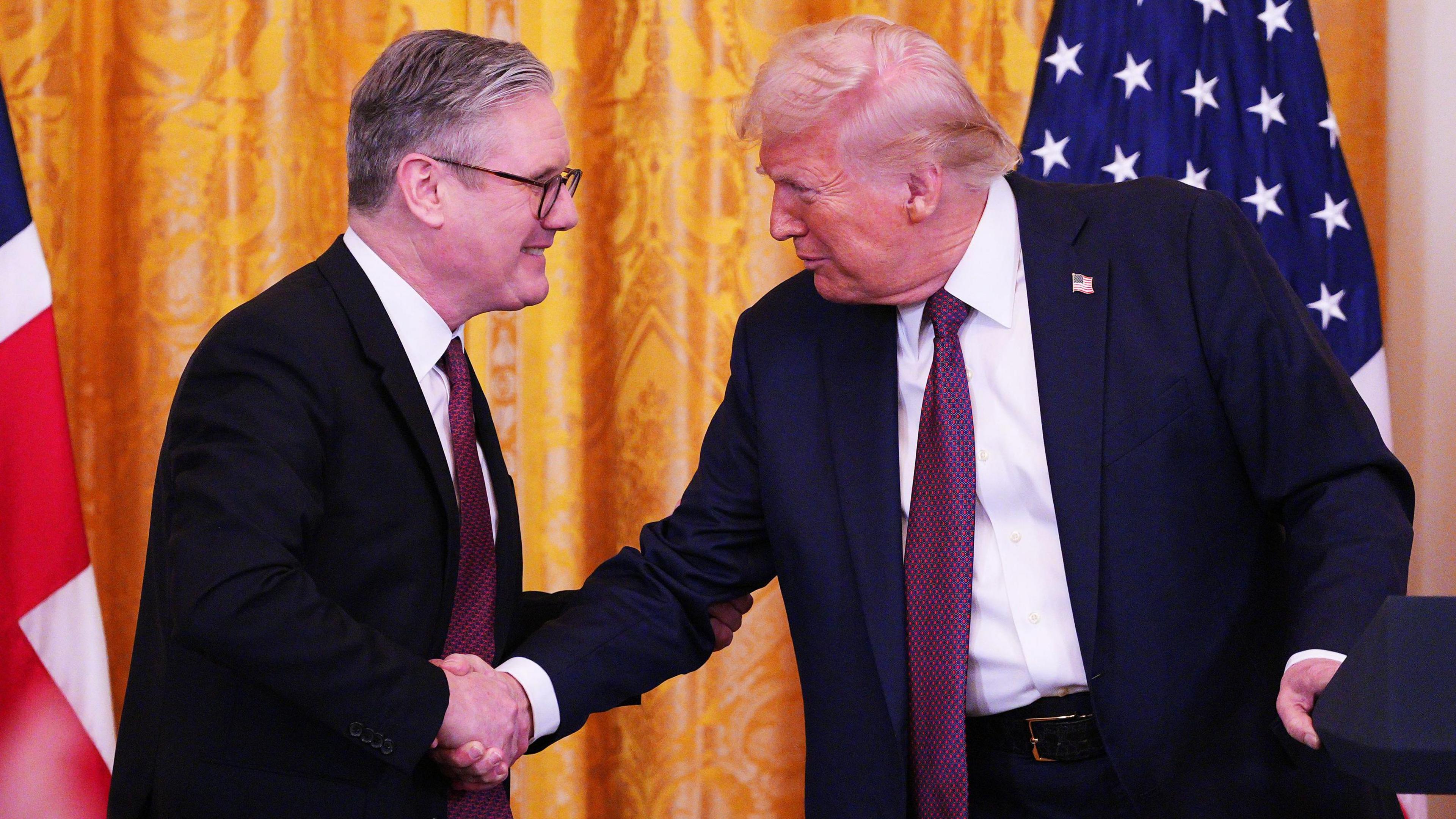 US President Donald Trump and Prime Minister Sir Keir Starmer shake hands during a press conference at the White House in February 2025. Behind them is a yellow curtain behind a Union Jack and US flag.