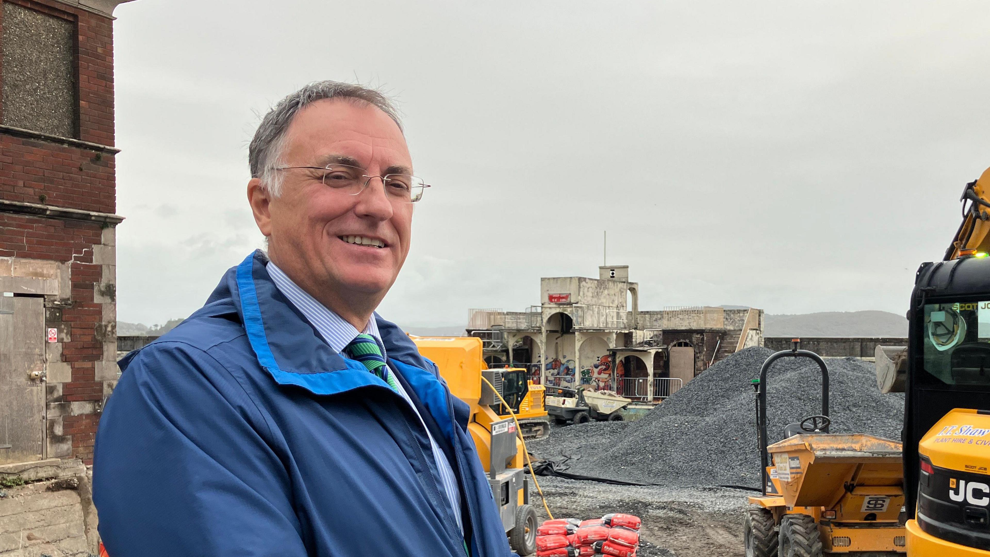 Jonathan Brook is standing on a construction site and smiling at the camera. He has short grey/black hair and is wearing glasses and a blur waterproof jacket.