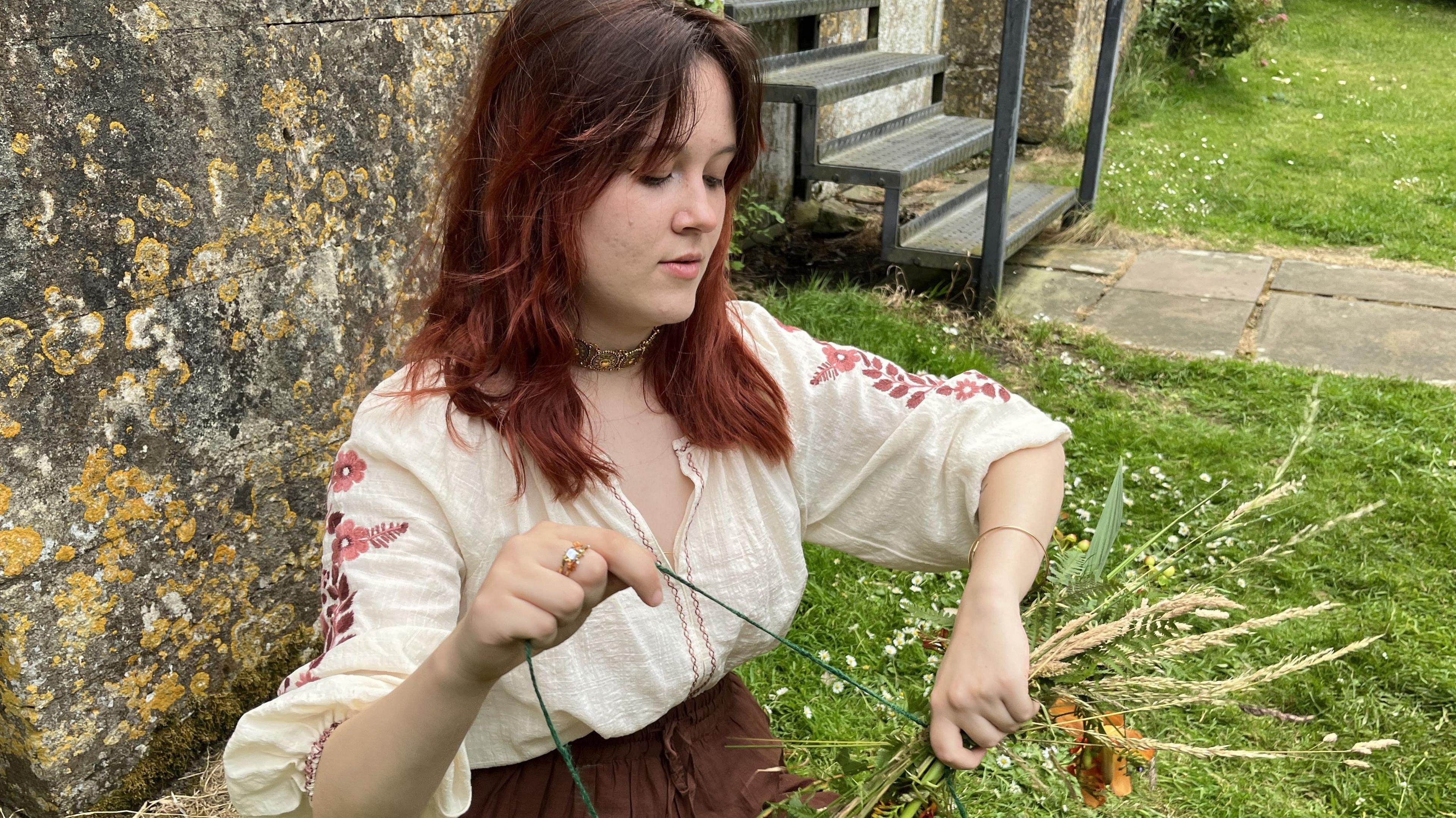 A Ukrainian refugee in traditional dress, is sitting in front of Malmesbury Abbey making a flower crown.