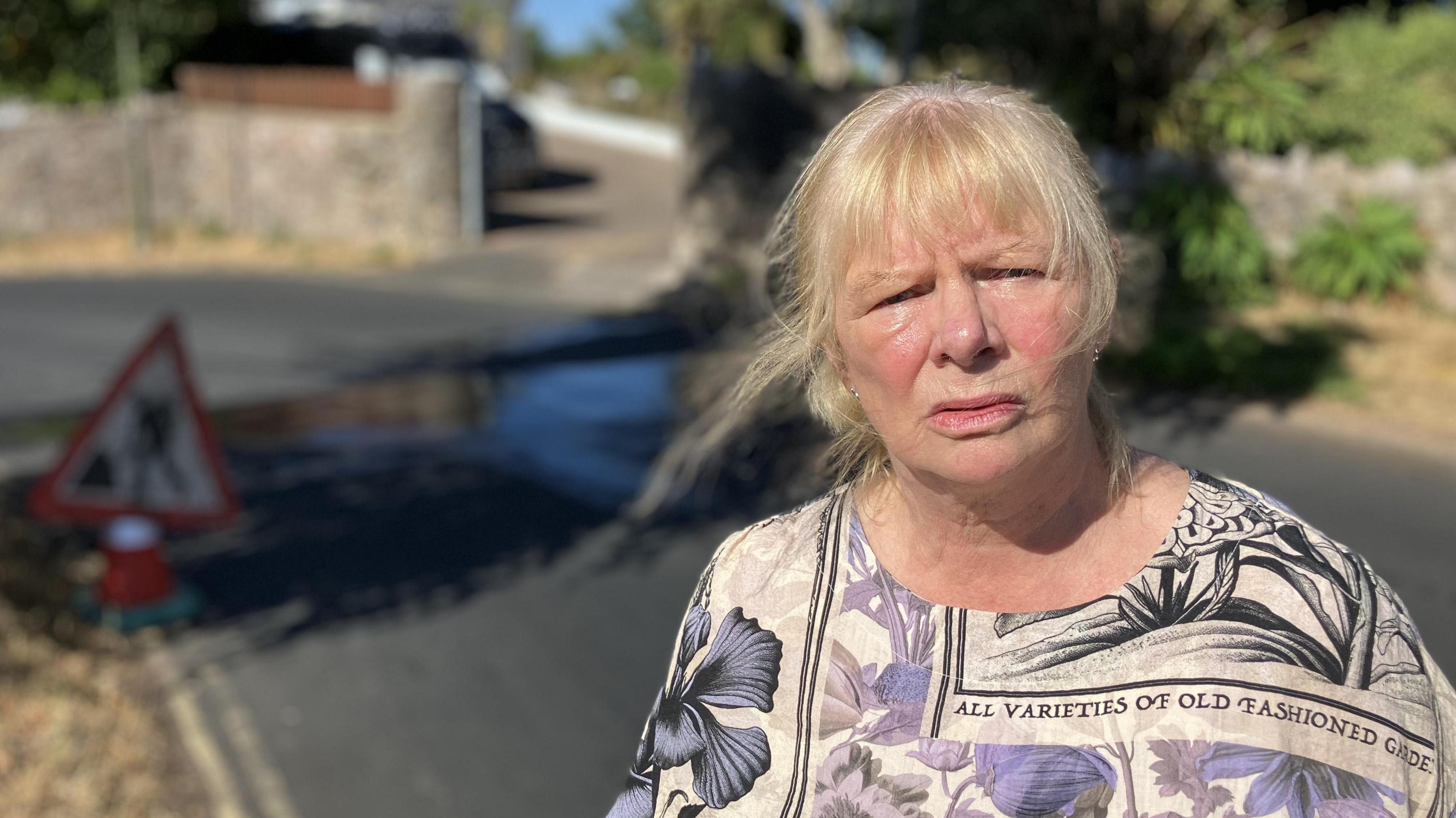 A woman stands by a water leak in Victoria Road in Brixham. There is water running down the road in the background from a leak on the verge. There is also a sign at the side of the road signifying roadworks. 