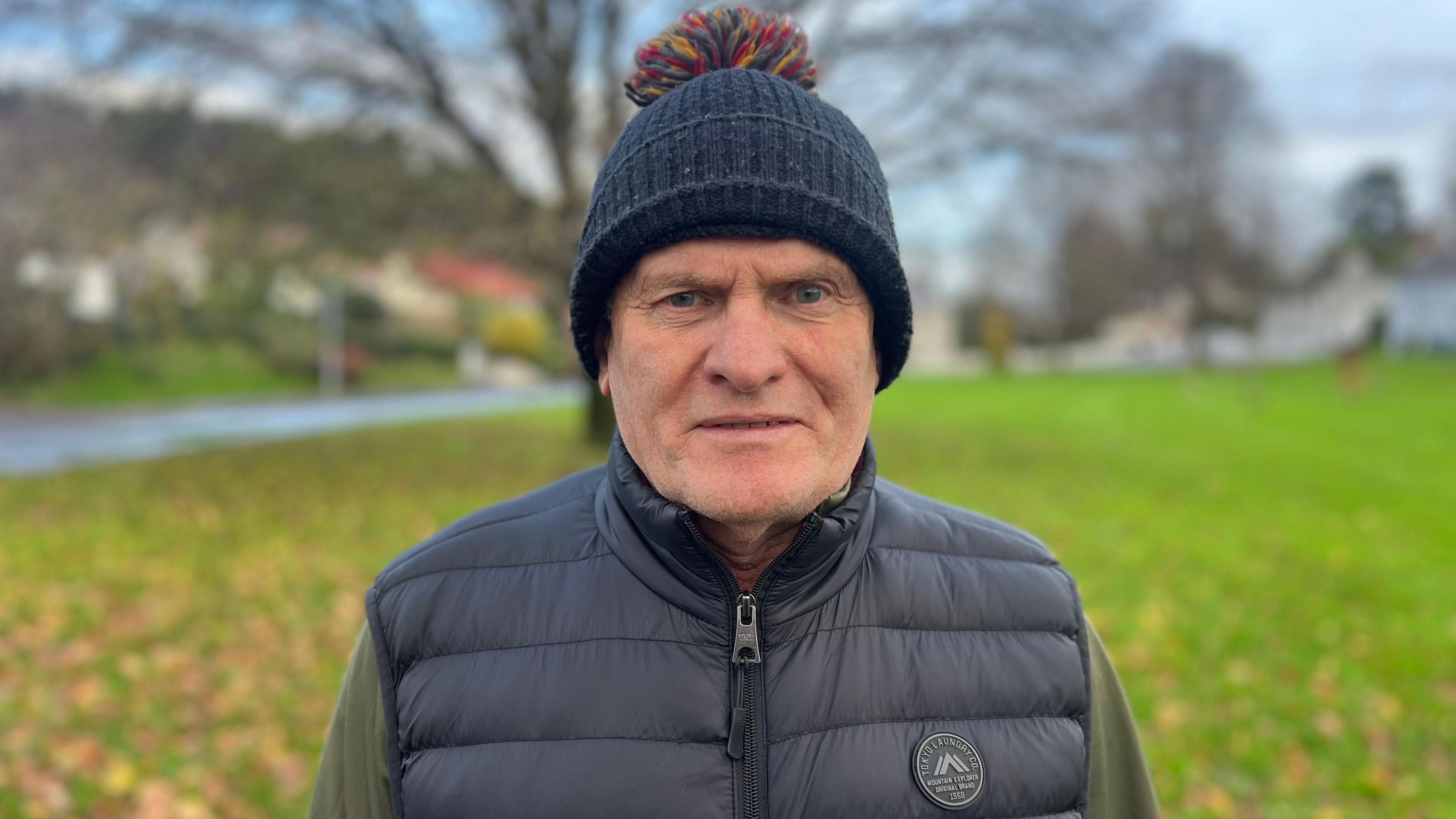 Trevor Love is standing outside with grass behind him. He's wearing a navy bobble hat and a navy body warmer with a green top underneath. The trees behind him are bare and leaves are on the grass.