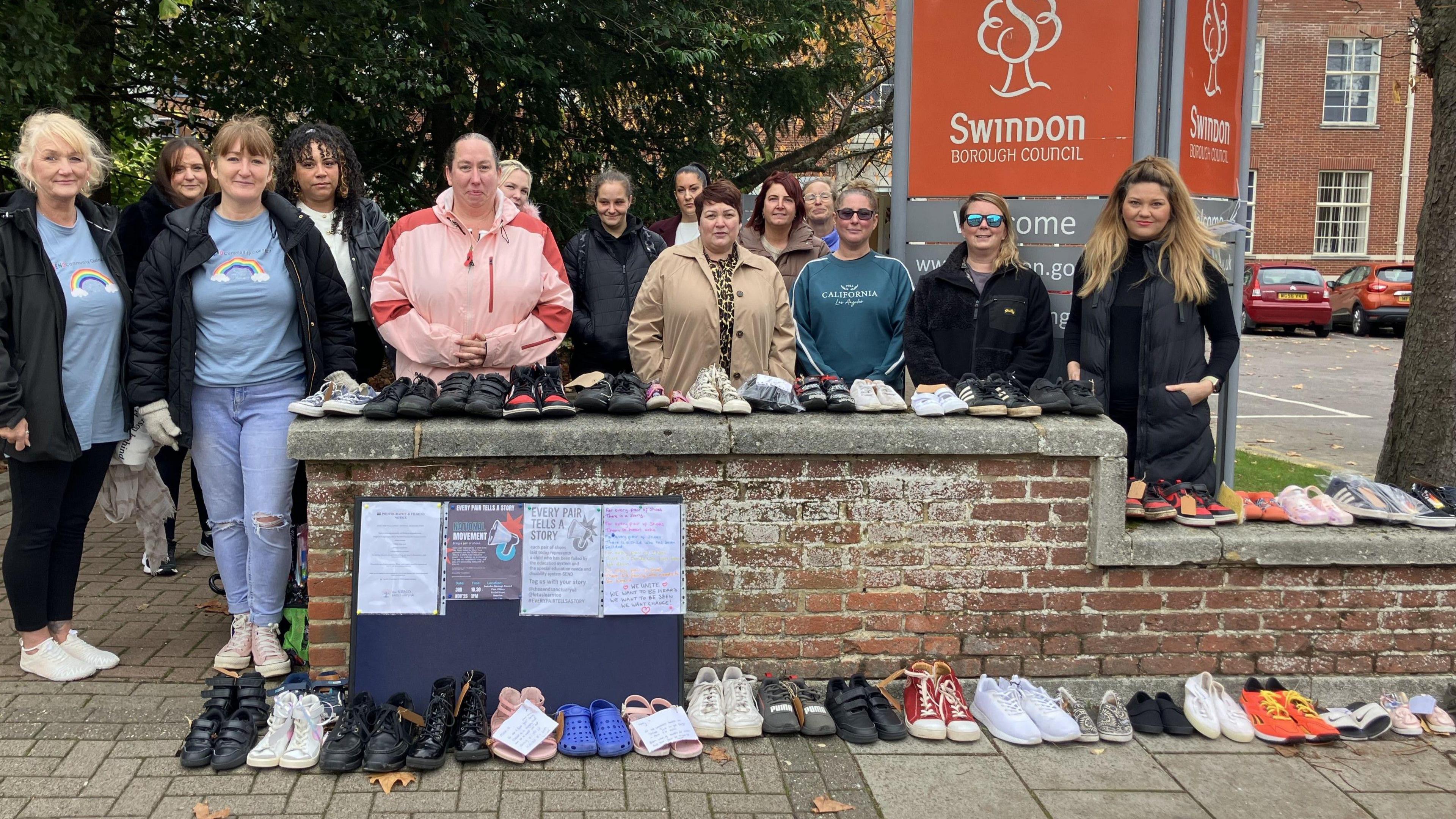 A group of women are stood behind a wall which has rows of children's shoes lined up in pairs on it. Some of the shoes have a paper tag on them with a message. The are stood in front of a 'Swindon Borough Council' sign.