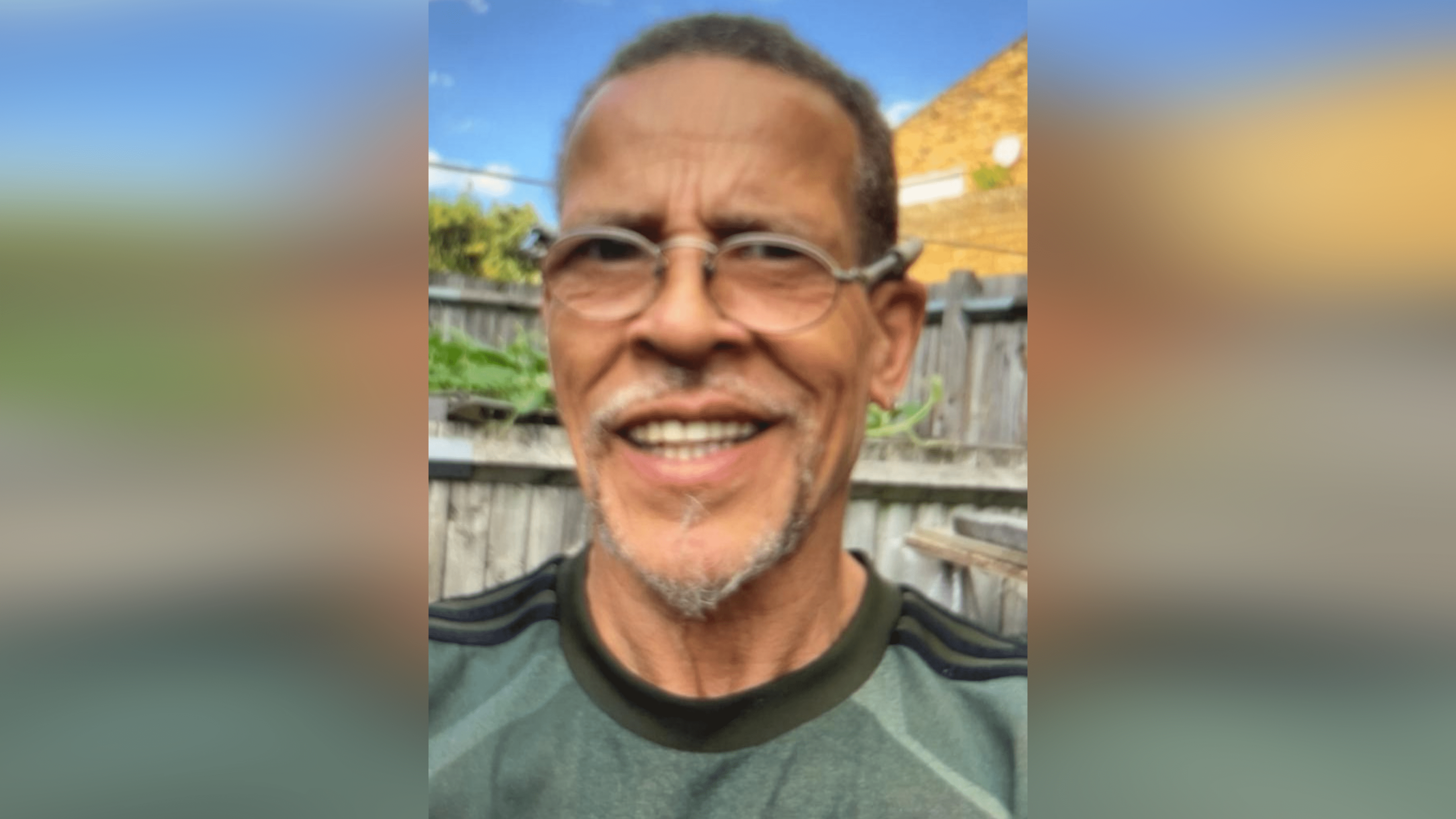 A man in a green t-shirt and glasses smiles at the camera. He stands outside, behind him is a wooden fence. 