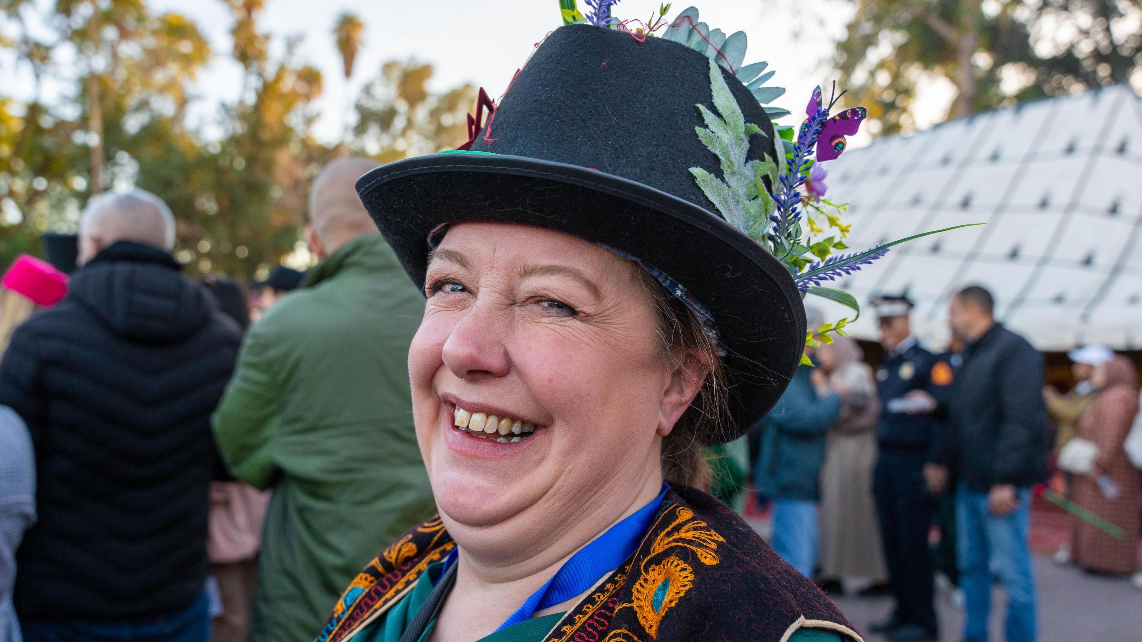 Veronica Chambers smiles at the camera. She wears a black hat with some flowers on it and the shoulders/lapels of her black jacket have orange and blue embroidery on them.