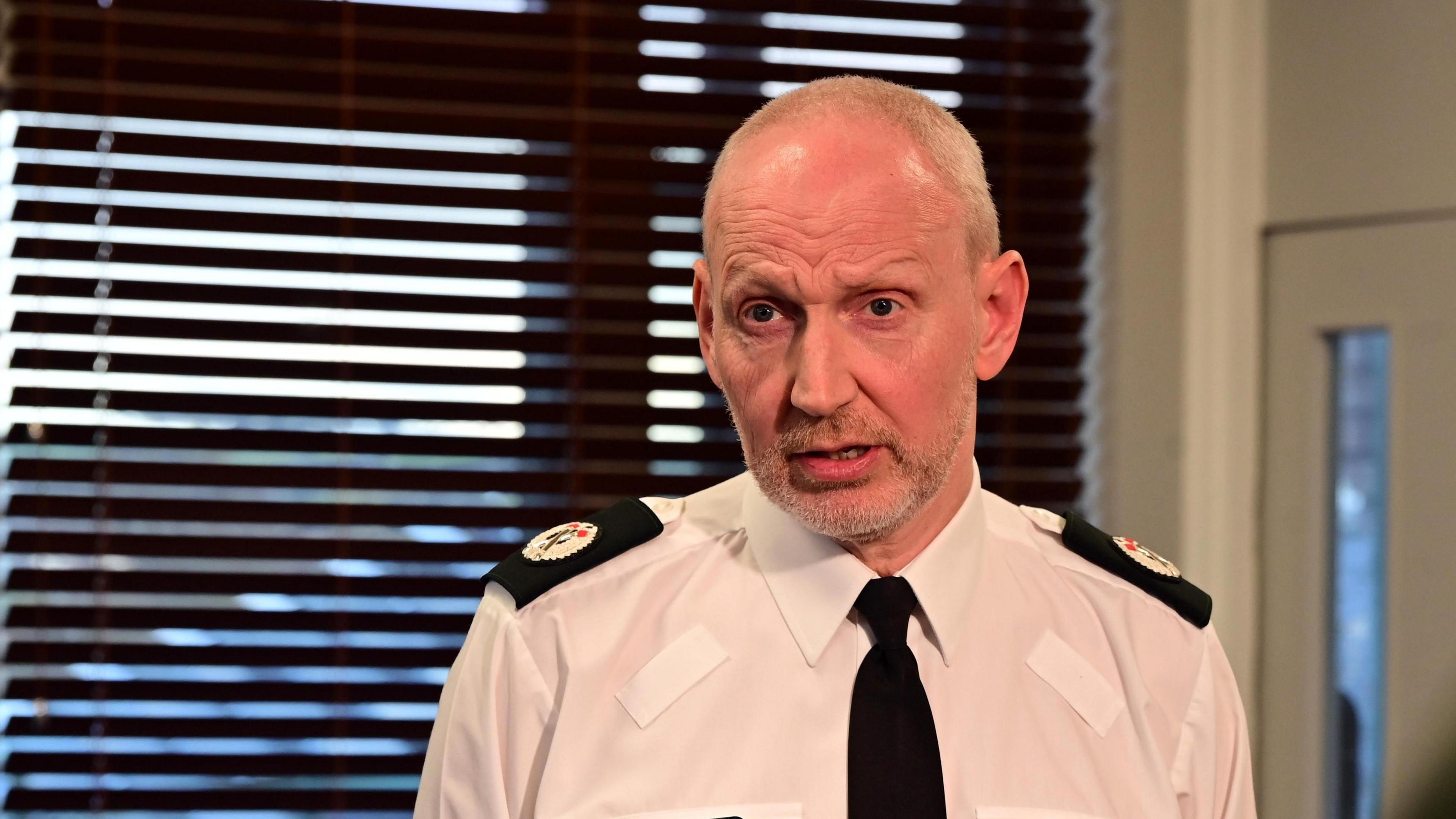 Assistant Chief Constable Davy Beck. He has shirt white hair, wearing a white shirt, black tie and black police shoulder patches. He is standing in front of a window with dark venetian blinds. 