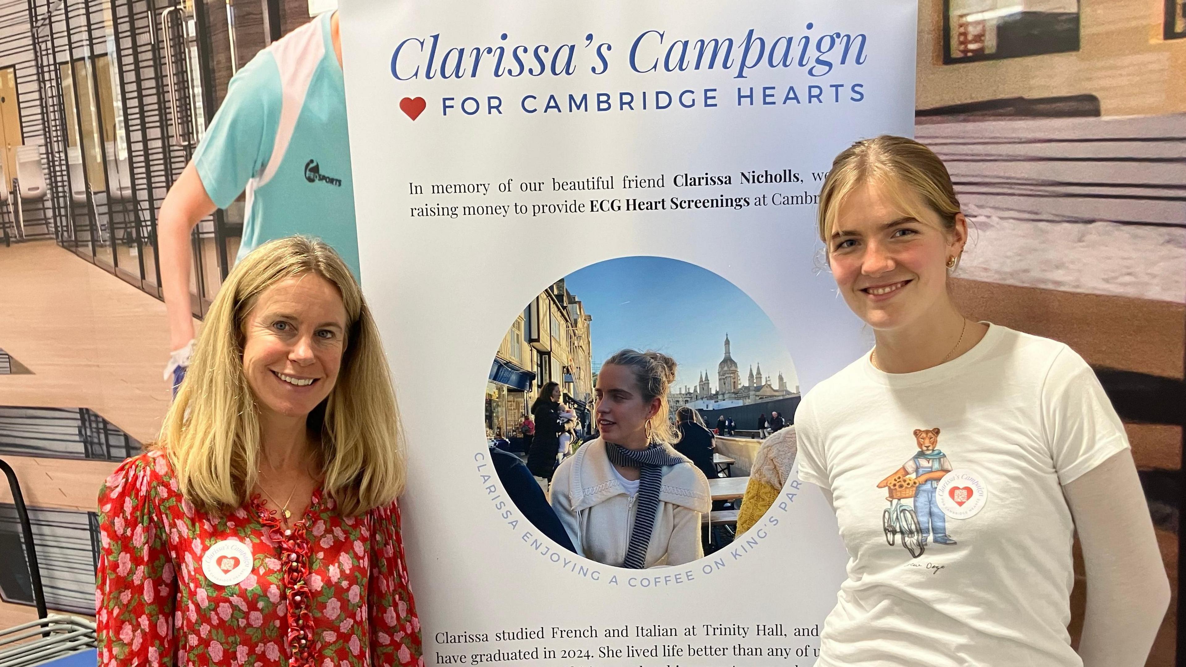 Two light-haired women - one, on the left, wearing a red and pink top, one, on the right, wearing a cream t-shirt bearing a teddy bear image - standing either side of a "Clarissa's Campaign for Cambridge Hearts" poster 