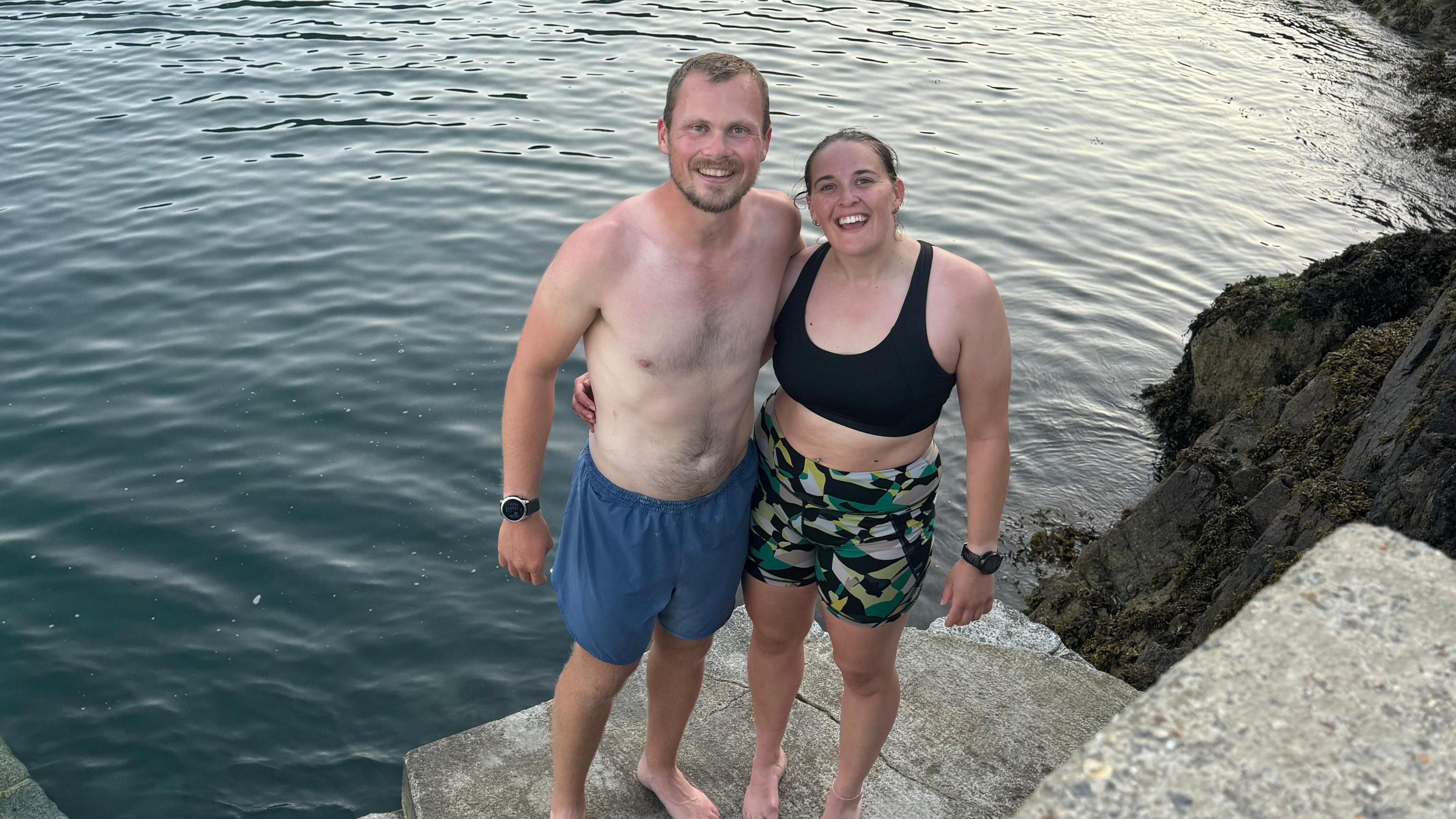 Matt Holland and Ellie Tanguy stood next to each other in front of one of the bays in Guernsey, they are wearing swimming gear and are about to get in the water.