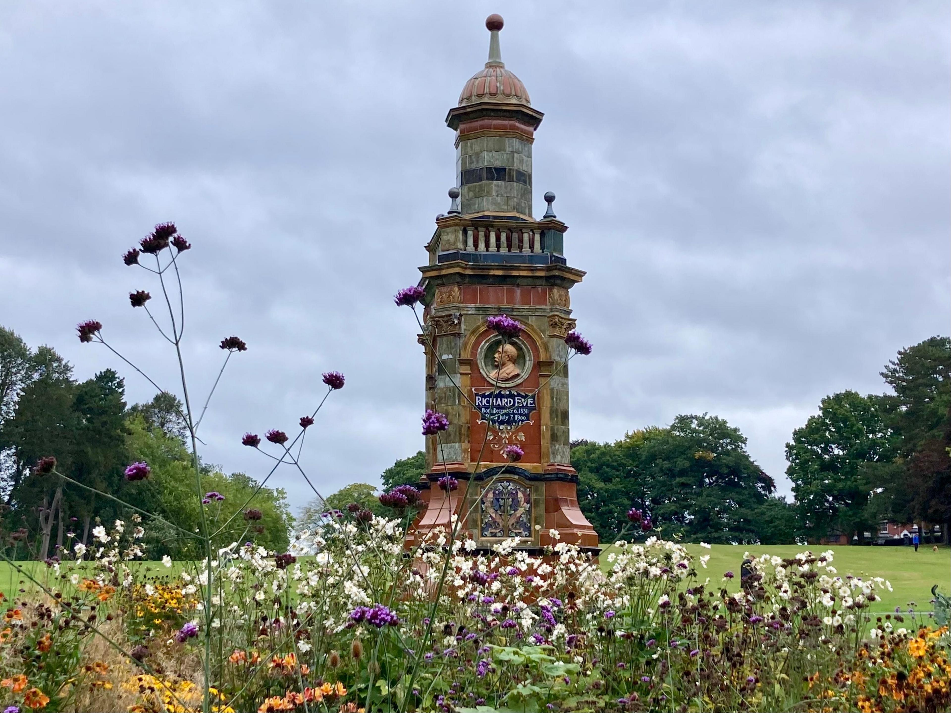 A memorial tower decorated with brown and grey ceramic tiles and a relief bust of Richard Eve above a memorial inscription in white on blue. In the foreground, purple, white and yellow flowers spring from a bed. 