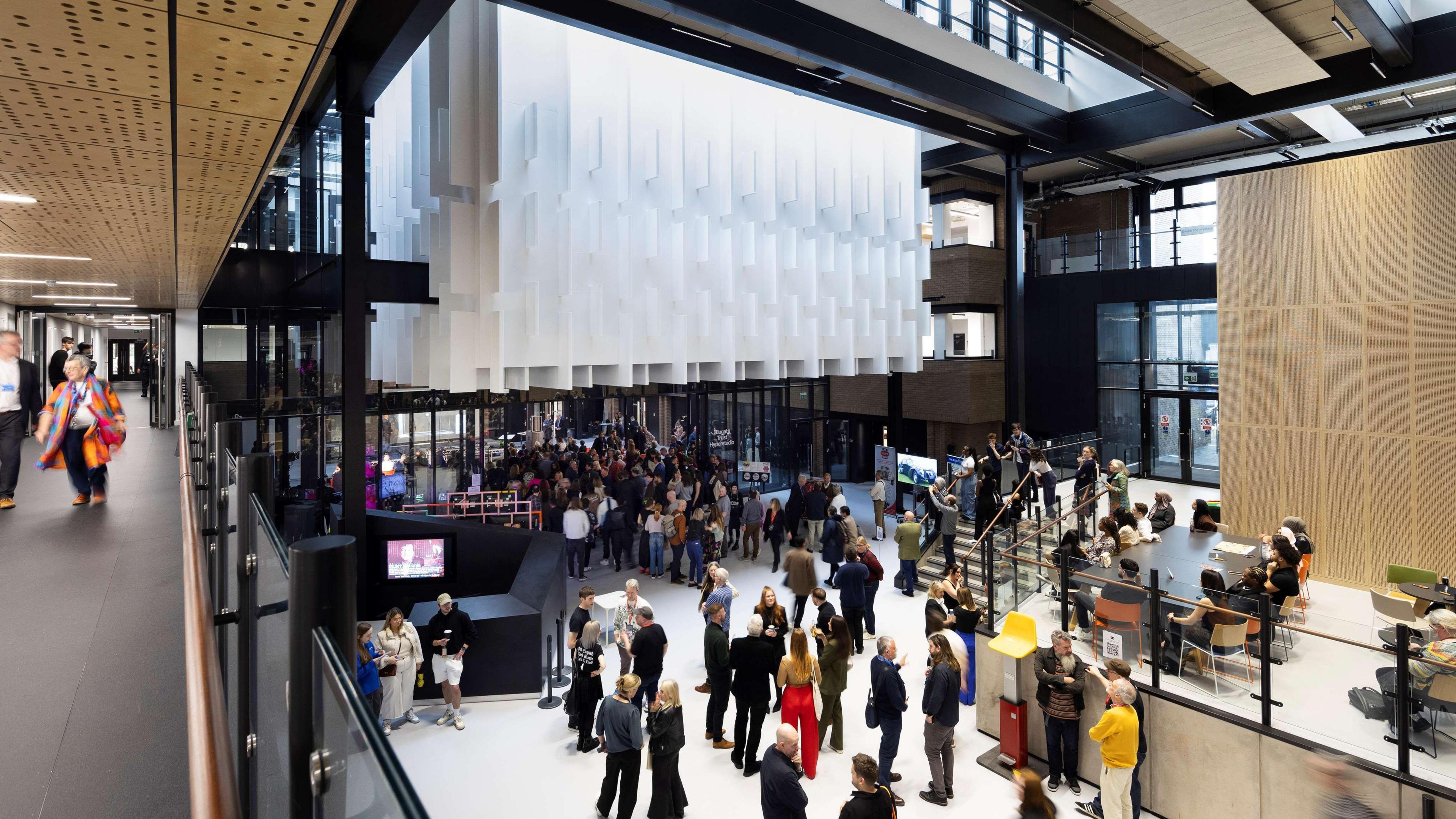 This is an image of a large open plan space filled with people. There is a large white screen hanging from the wall above. There are people sitting around tables on a mezzanine level to the right and a black railing above the ground floor to the left. 