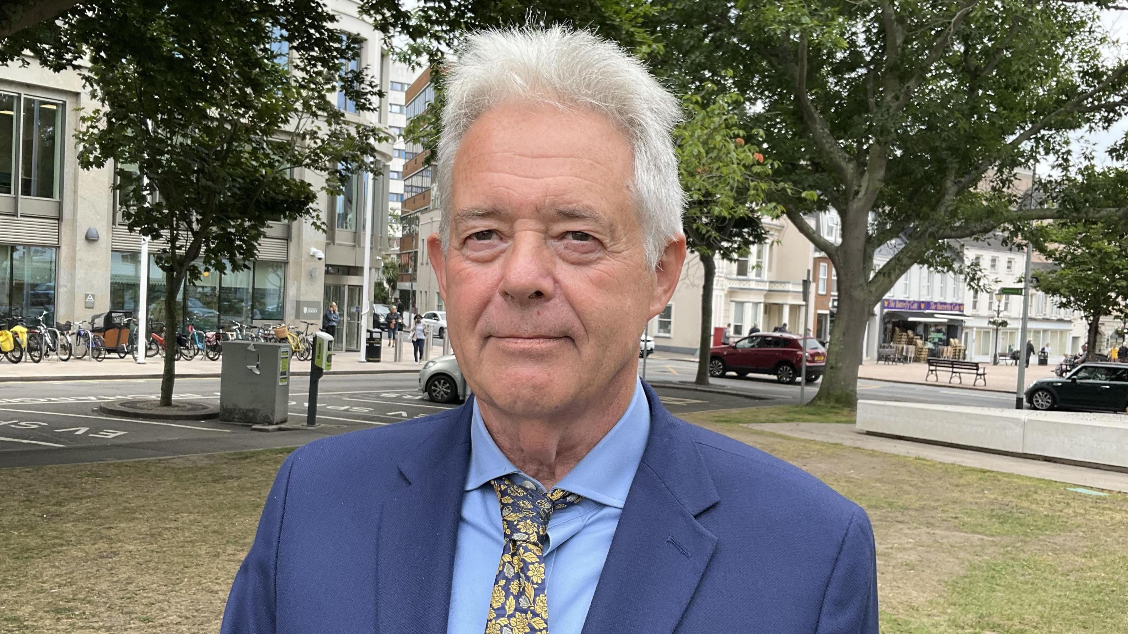 Barry Freeman in a blue suit, blue shirt and flowery tie. He is stood in Parade gardens, with trees and roads visible in the background.