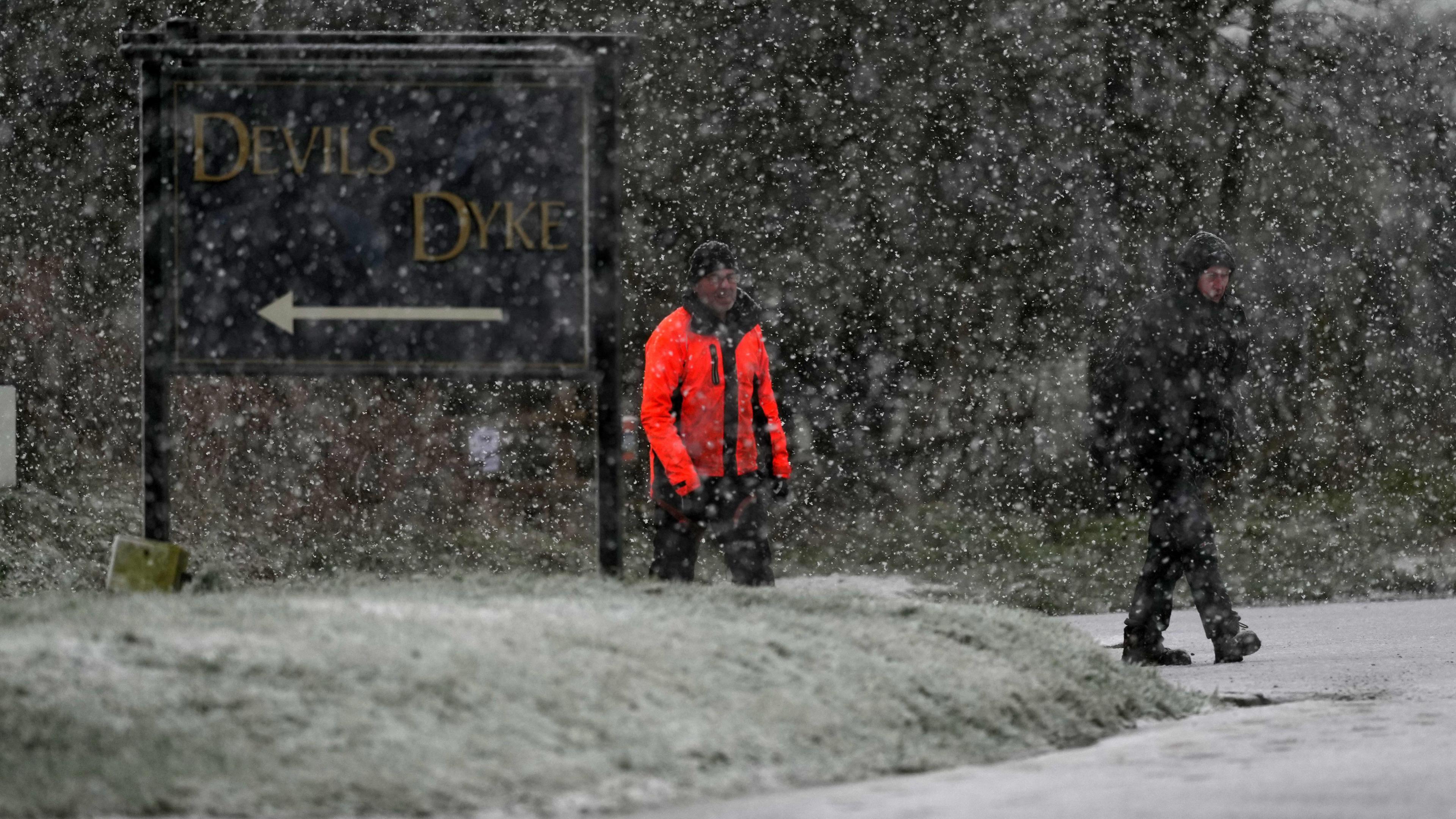 Two men walk past a sign saying Devil's Dyke while snow falls