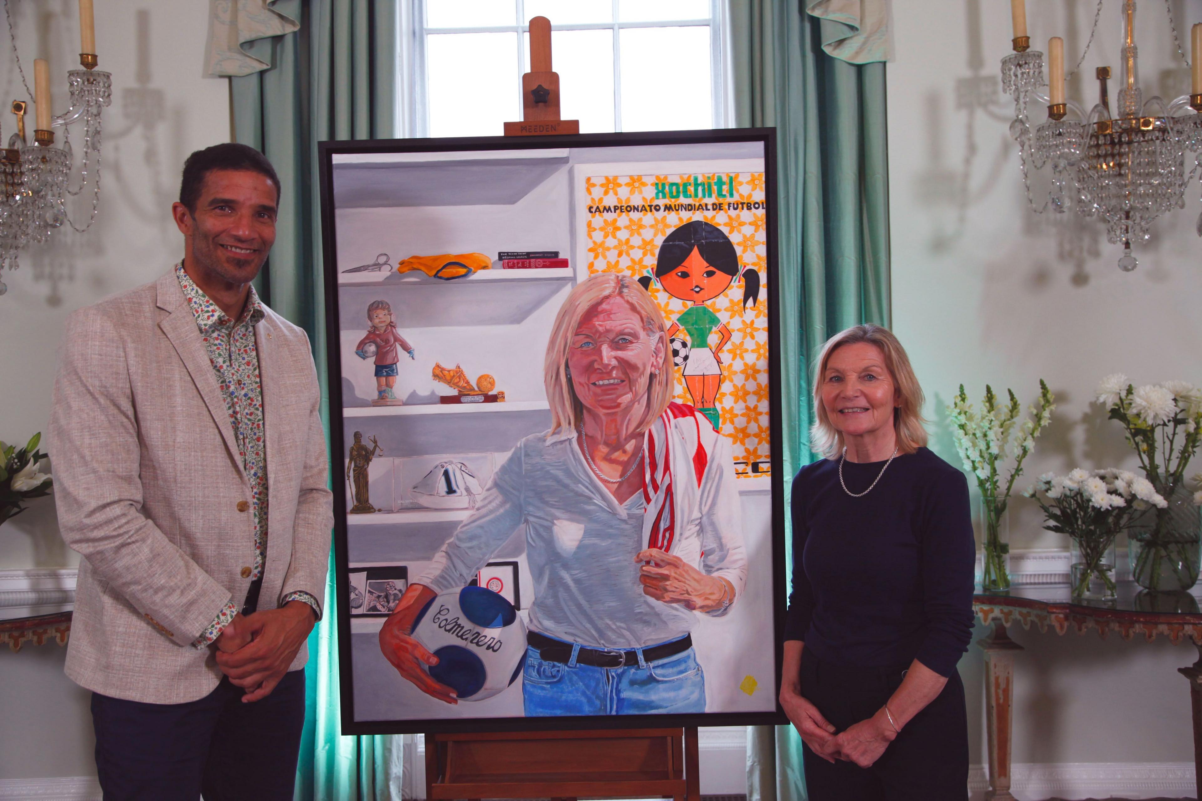 David James, in linen blazer and flower-print shirt, stands with Gill Sayell, in a black shirt, next to the former goalkeeper's portrait of Sayell, who is depicted in blue jeans, with a white shirt, holding a football and surrounded by football memorabilia. 