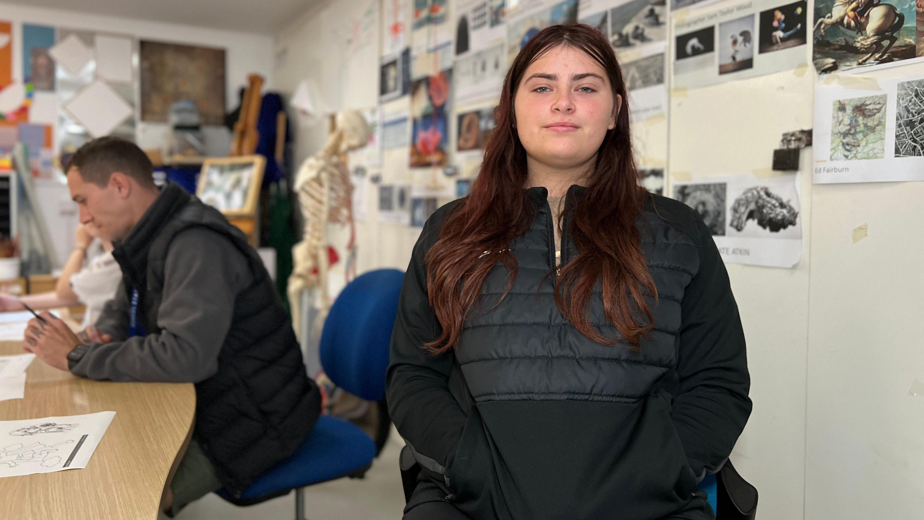 Tiarni is looking at the camera and wears a black top with her hands in her pockets. She's in an art classroom with drawings on the walls and a skelton in the background. Behind her to her left is a teacher holding a pencil looking at paper. Tiarni has long dark hair.