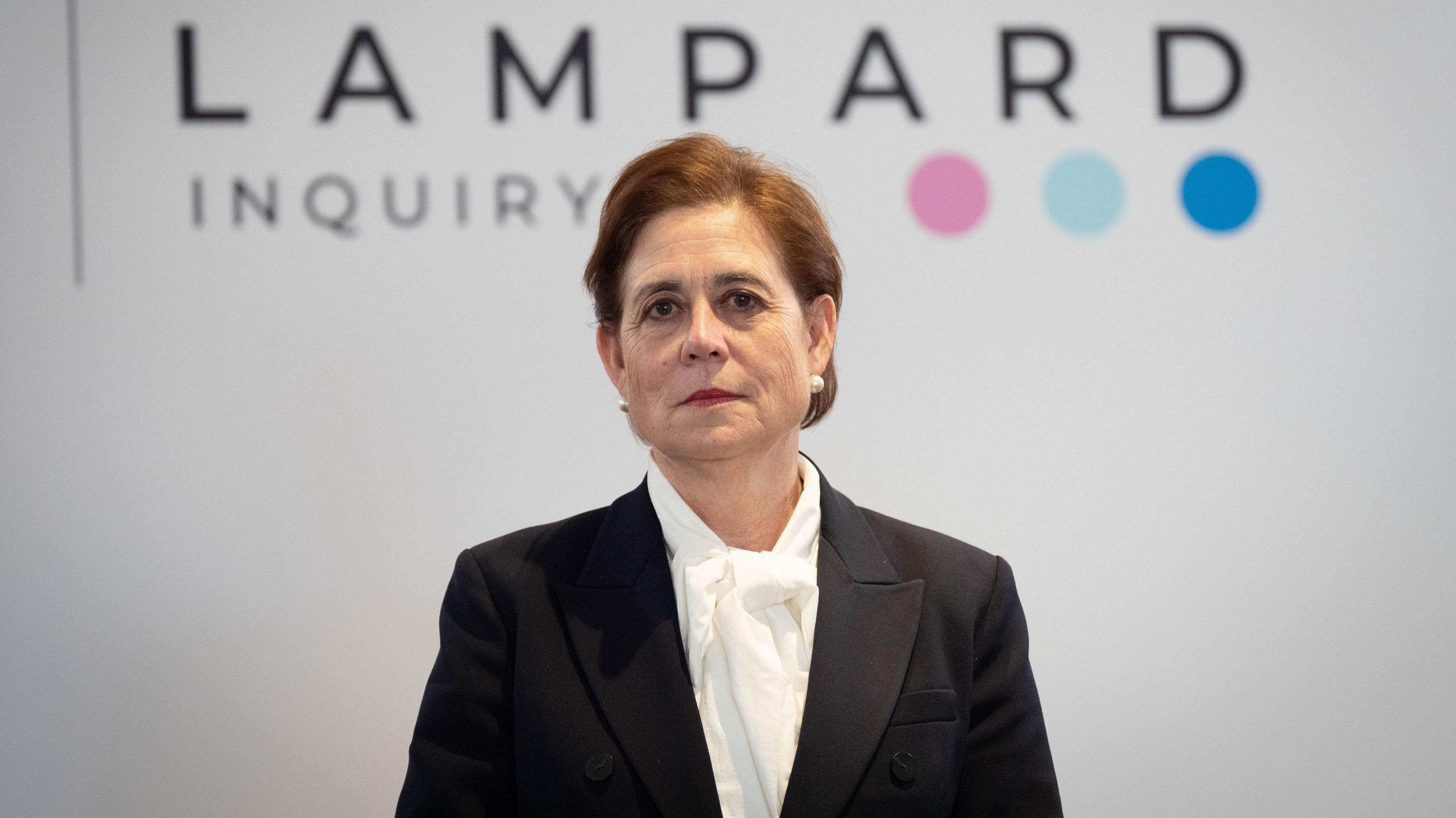 Baroness Lampard stands in front of a white background with the words Lampard Inquiry written on them. She has dark brown hair with a side parting, large white pearl stud earrings and has dark red lipstick. She is wearing a black blazer jacket and a white blouse.
