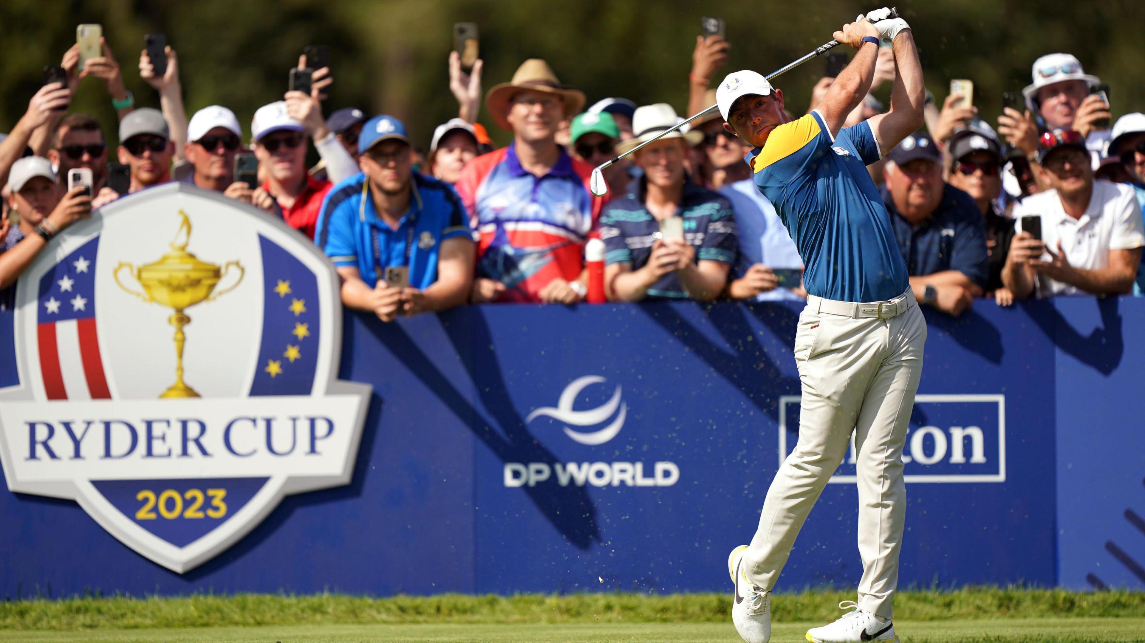 Action shot of Rory McIlroy playing a tee shot at the 2023 Ryder Cup. He is wearing the Team Europe outfit of a blue and yellow shirt and cream trousers. A throng of excited-looking fans stand behind him, some of whom have camera phones in hand.