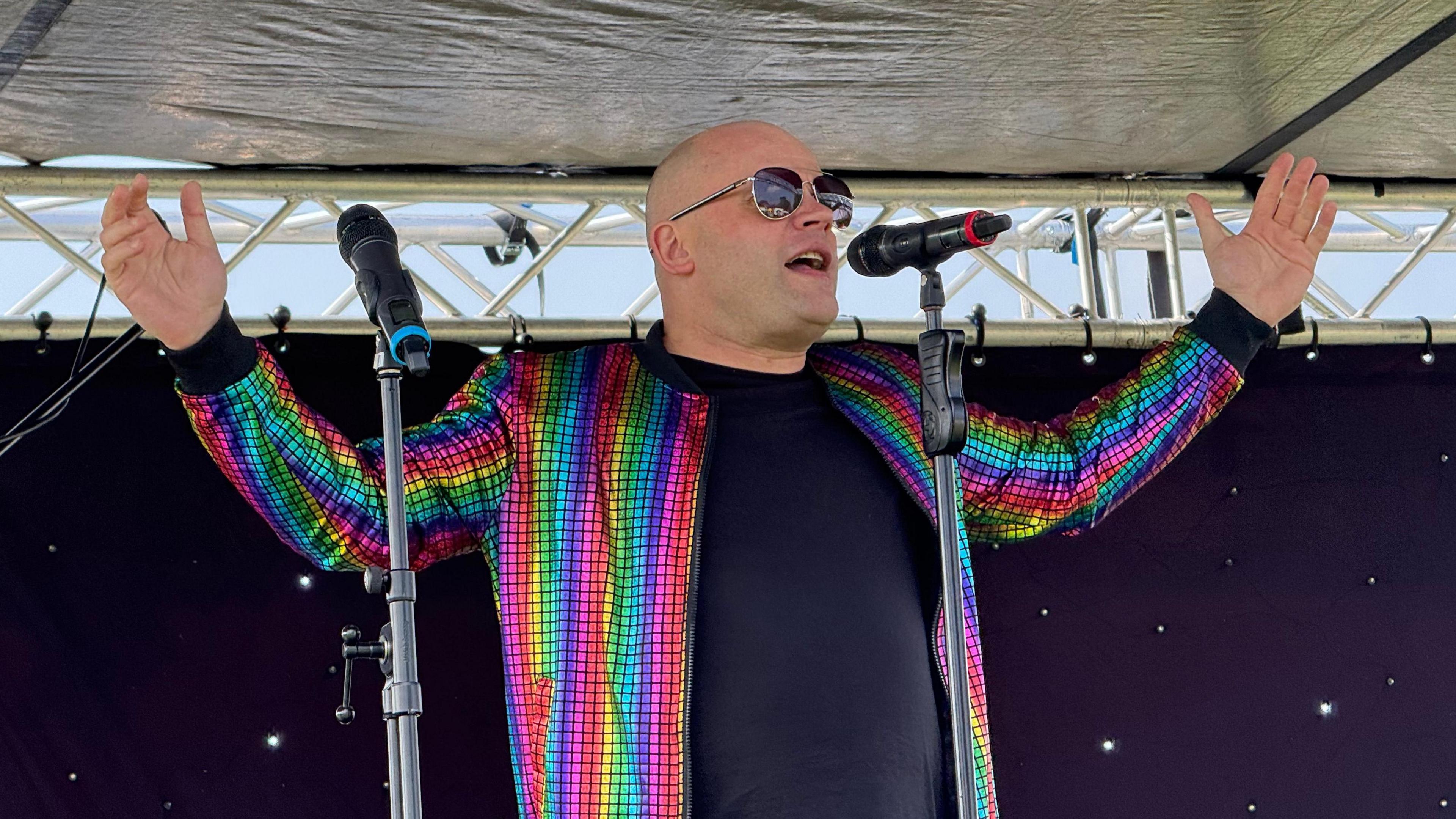 A man wearing sunglasses and a rainbow-coloured jacket with arms open is singing into a microphone.