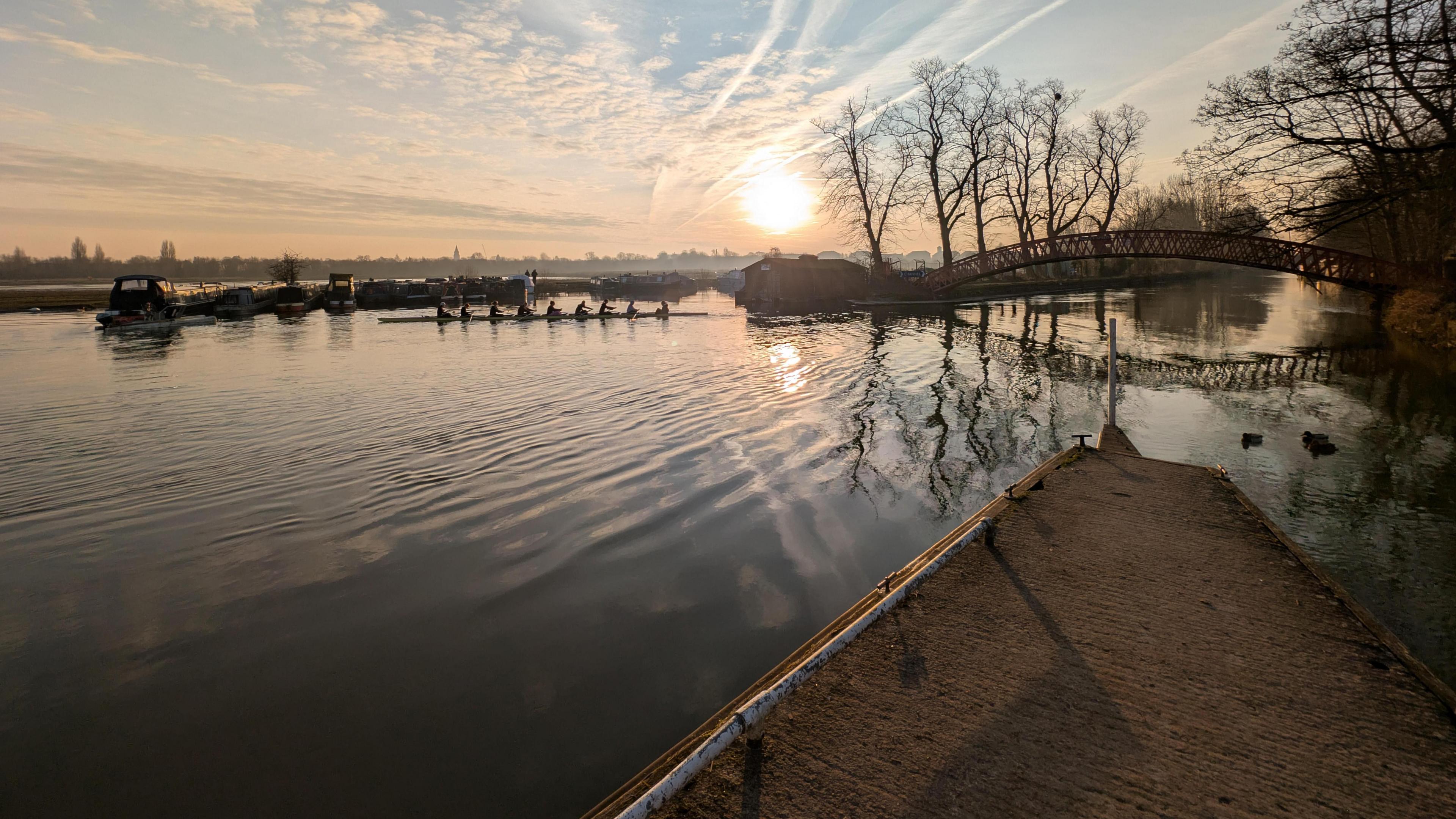 The sun glints off the water of the river. In the distance are narrowboats, a metal footbridge, and a boat with eight female rowers and what looks like a cox on the end.