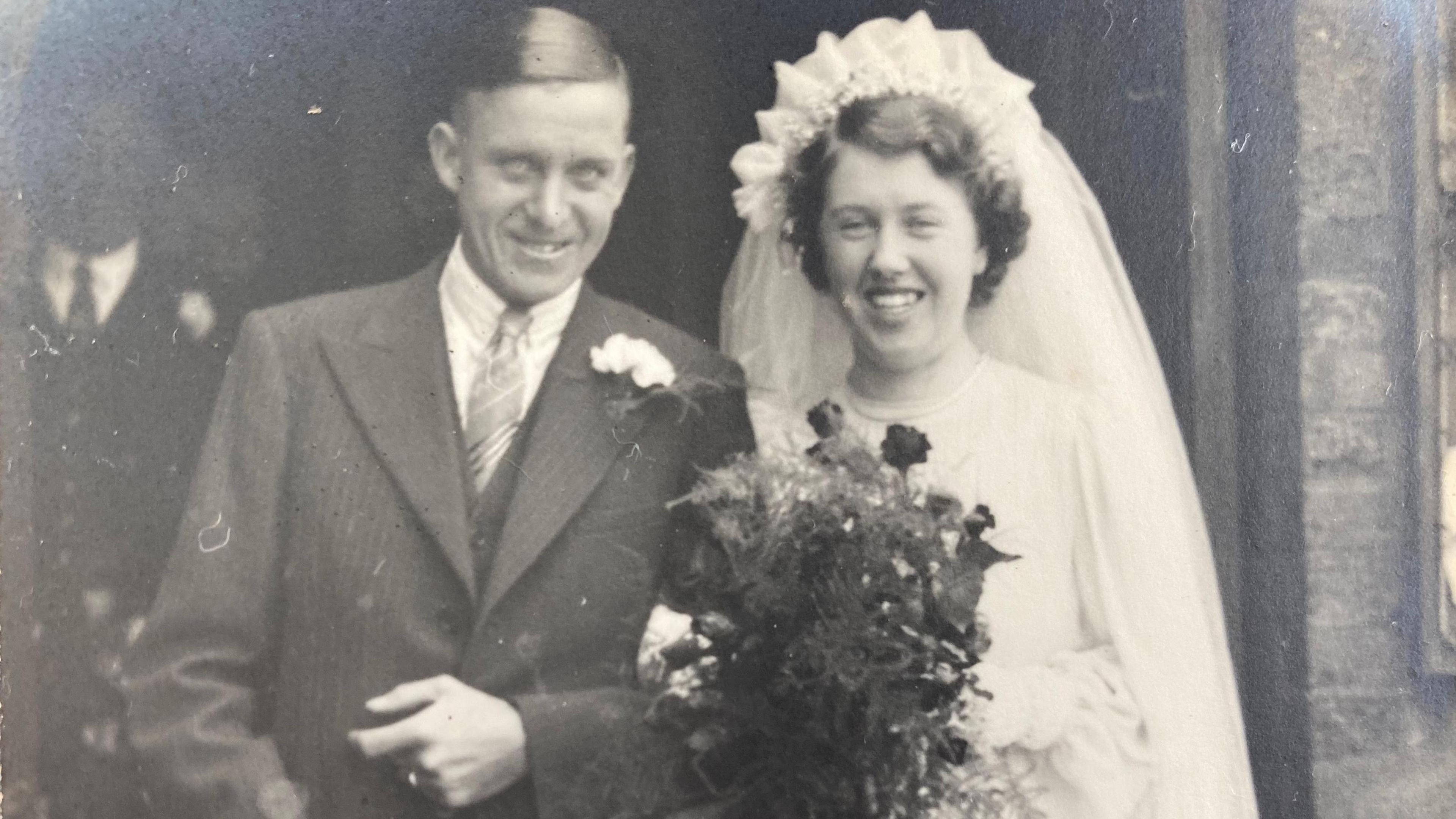 Leslie and Doreen on their wedding day, in a black and white image. He is wearing a suit, with a flower in his lapel, Doreen has a veil and head dress on, a white wedding dress and is holding a bouquet of flowers. 