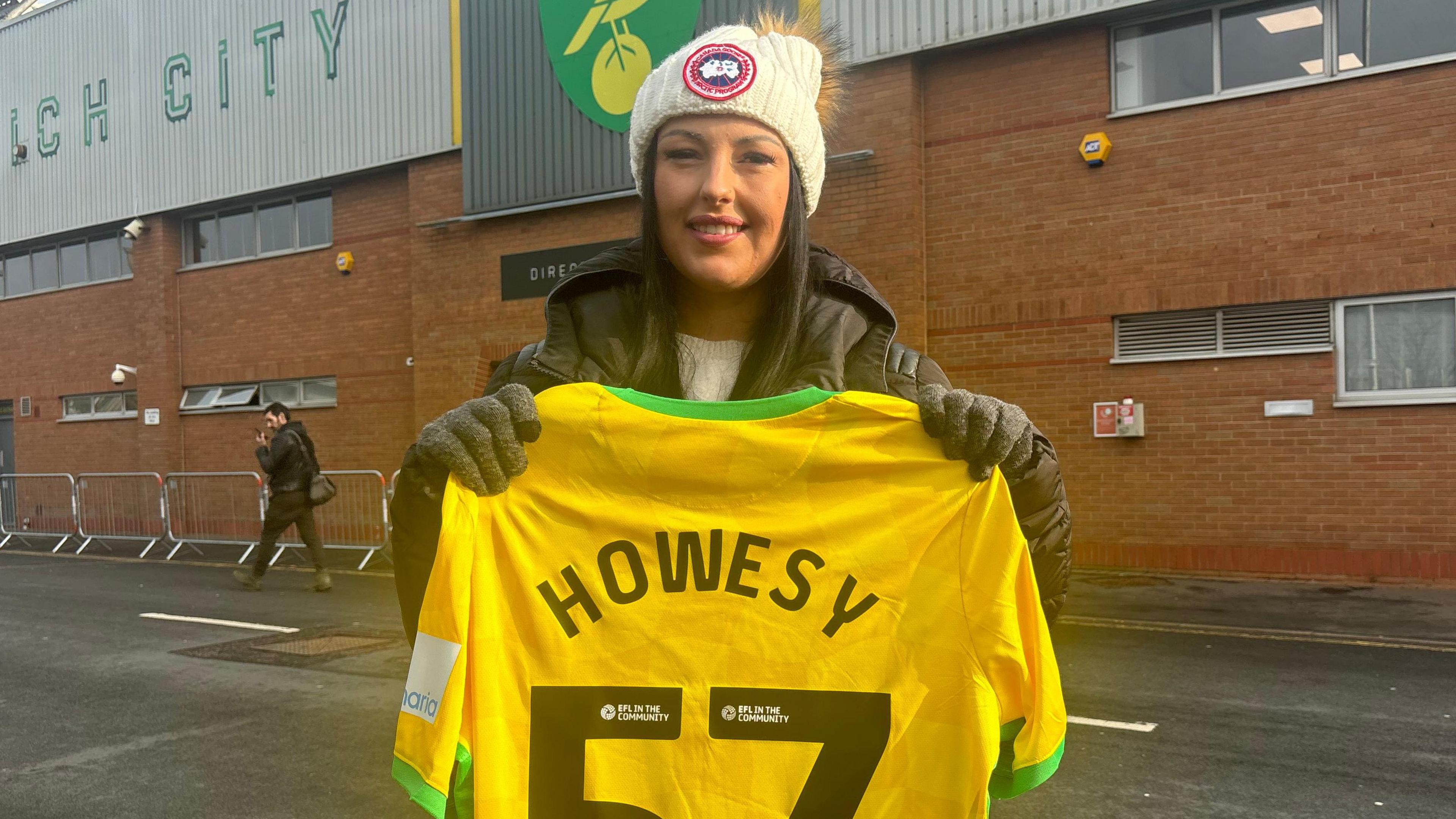 Livvy is seen standing in the middle of the picture with a black coat on, a white hat and is smiling at the camera. She is holding a Norwich City football shirt, which is yellow and green, which has "HOWESY" on the back above the number 57. 