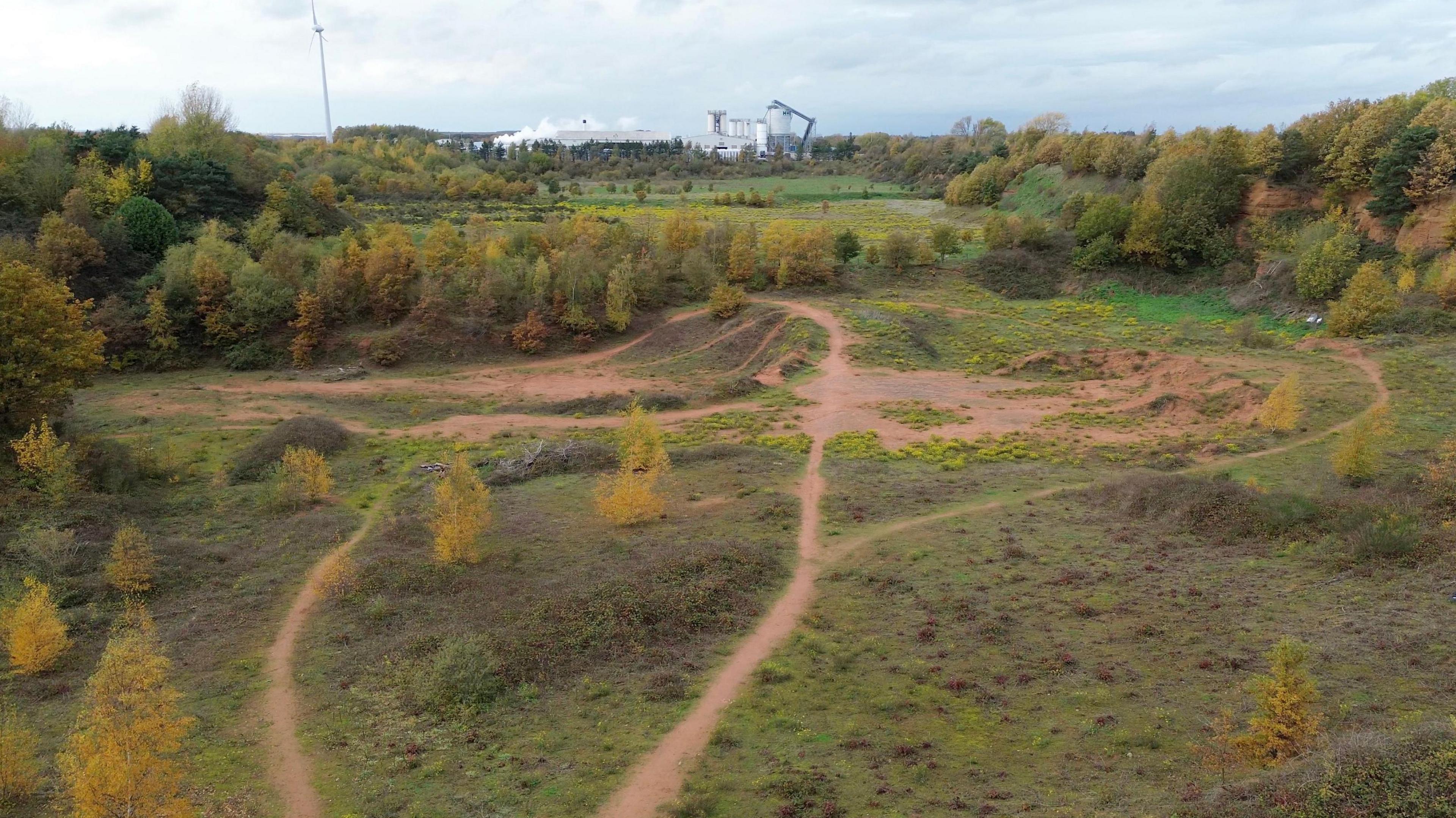 Footage from a drone shows the former quarry site. There overgrown scrubland surrounded by young trees. There is an industrial area in the background.