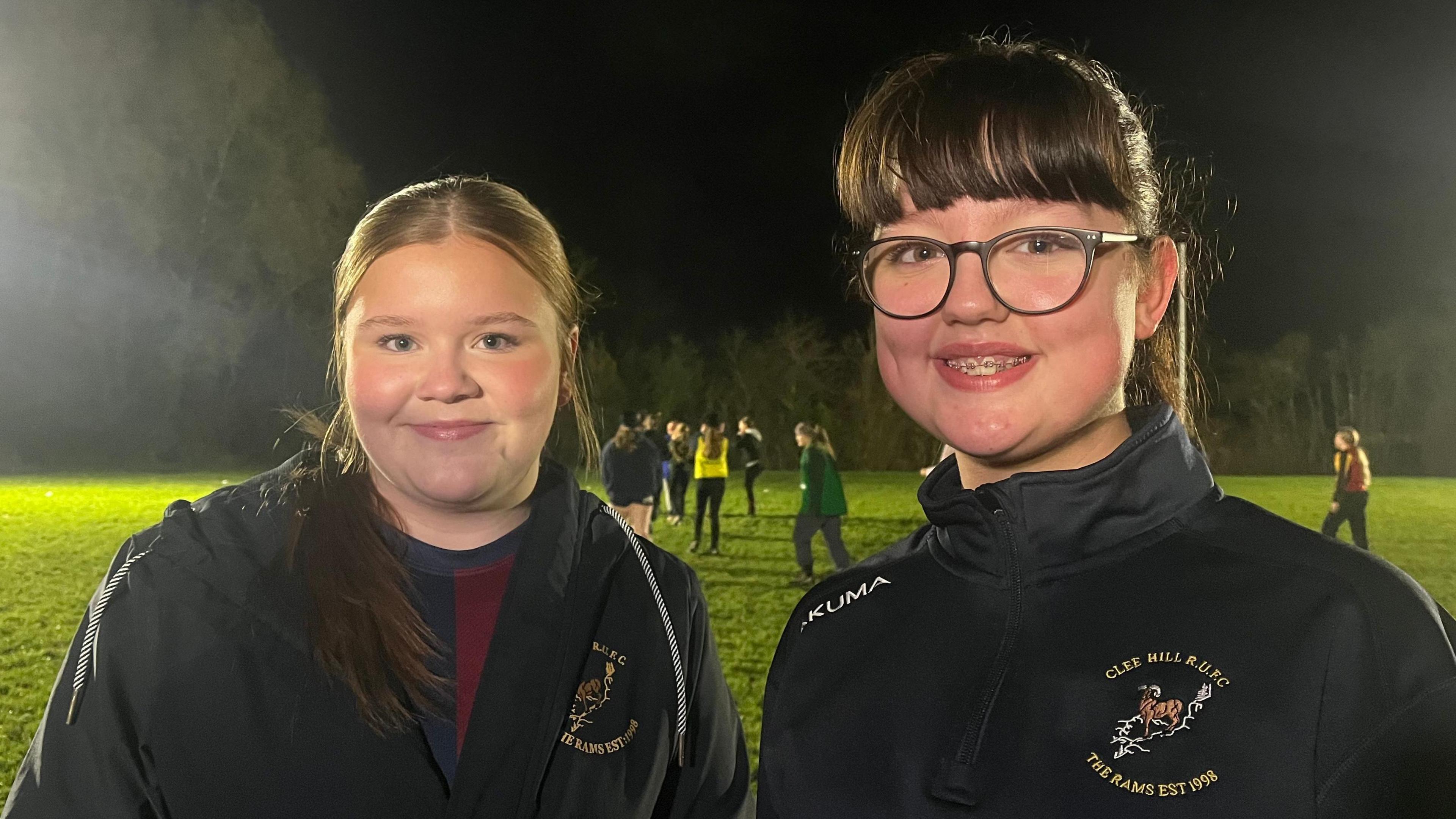 Eden and Jess stood side by side, both smiling and looking into the camera. They're stood on a flood-lit rugby pitch, which has green grass, a dark night sky, and is bordered by trees. Both girls are wearing black zip-up sports jackets, which have a logo on the chest that features a ram, and 'Clee Hill RUFC, The Rams Est 1998' written on it. 
