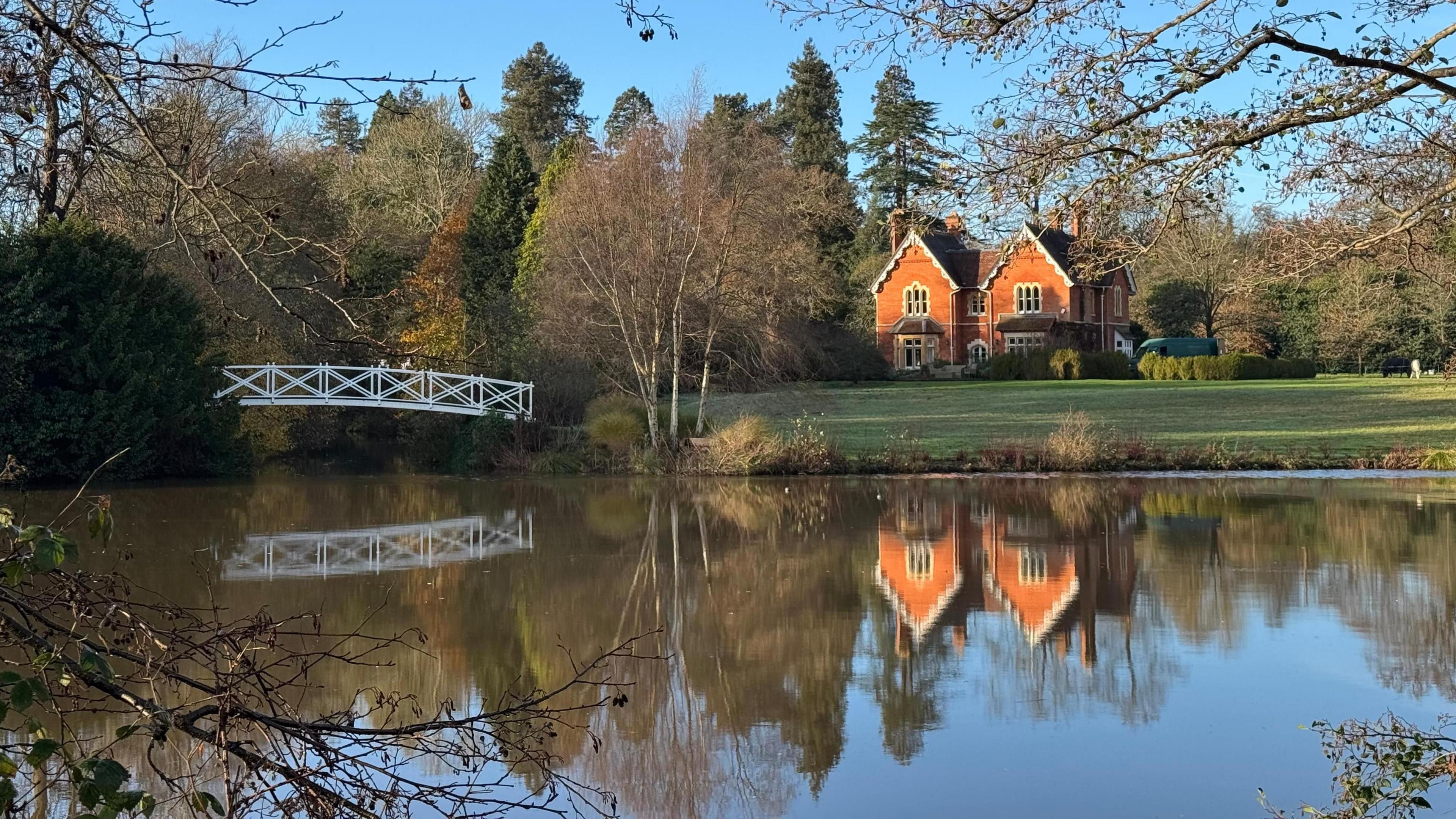 A red brick double-gabled property stands basking in winter sunshine. A well manicured lawn lays out before it leading to a still pond. A reflection of the house, trees and the blue sky are cast on its surface. To the left of the image is a small arching wooden bridge in white.