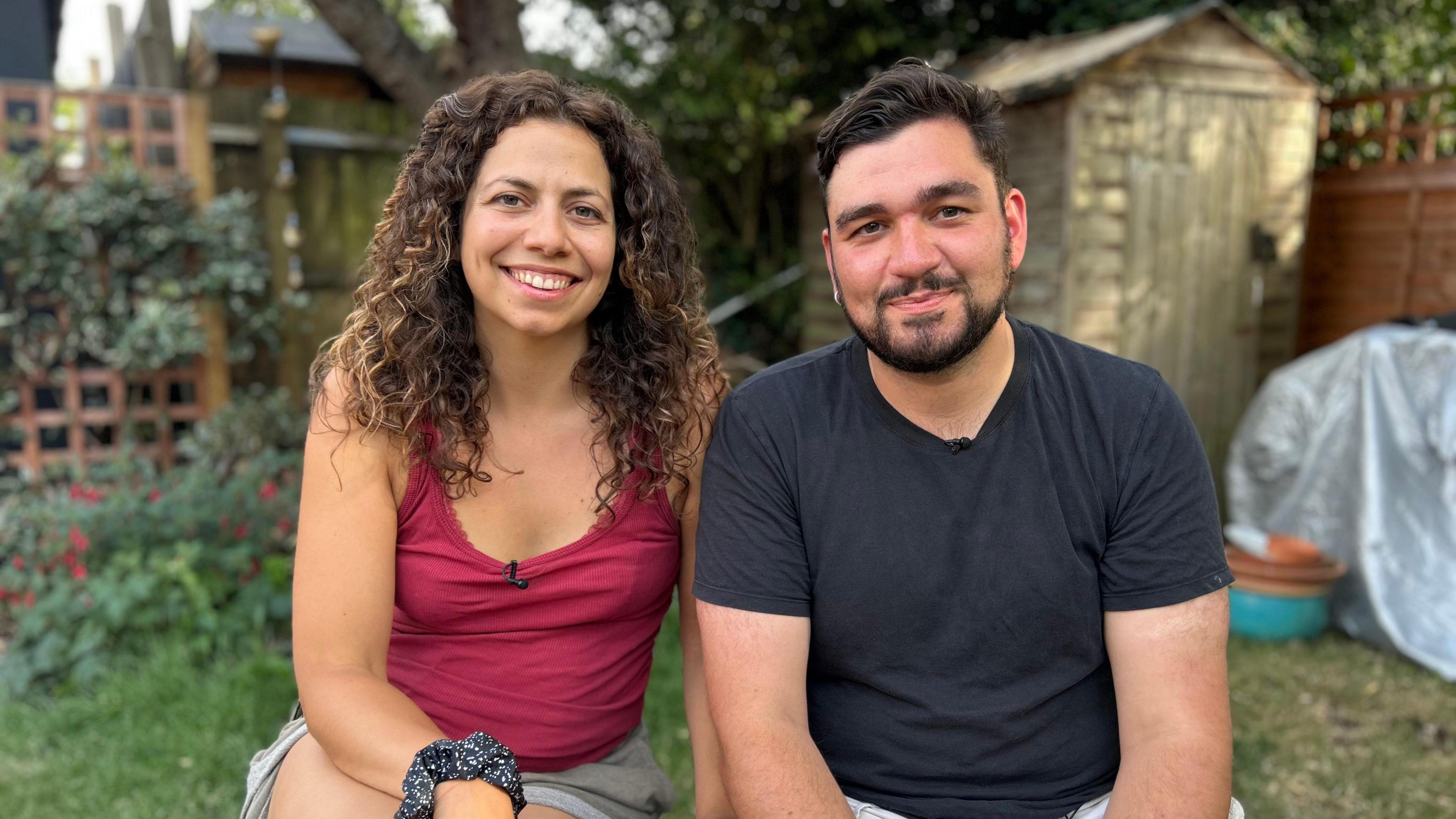 A couple in their thirties sit next to each other in the garden, smiling at the camera. The brunette man wear a dark t-shirt and the woman wears a maroon vest top.