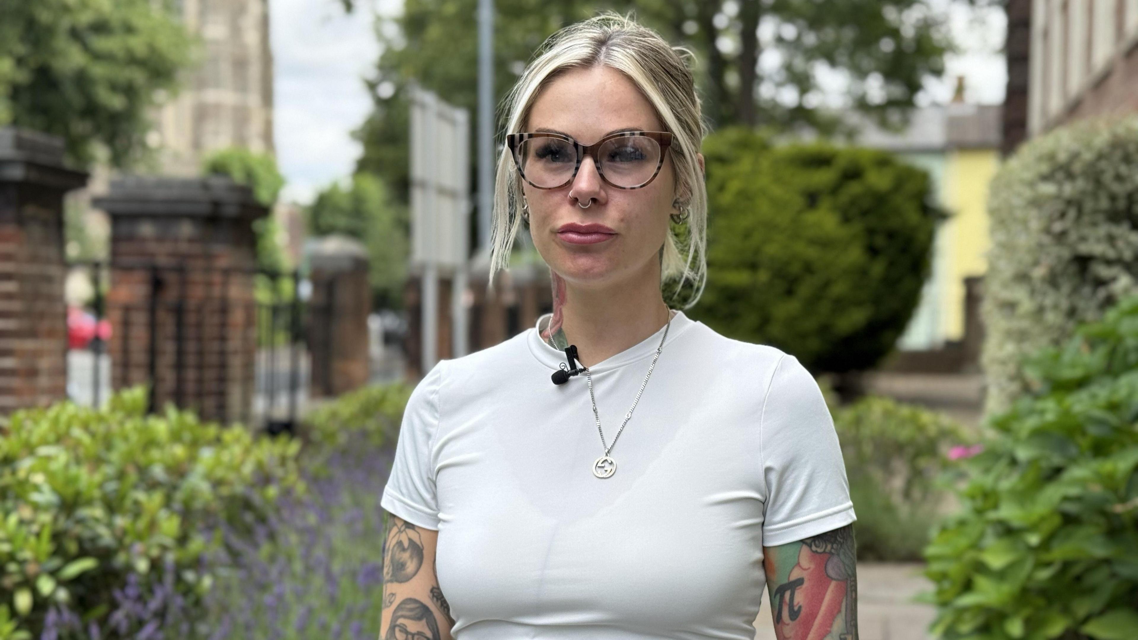 A woman with tattoos wearing a white-shirt looks into the camera.