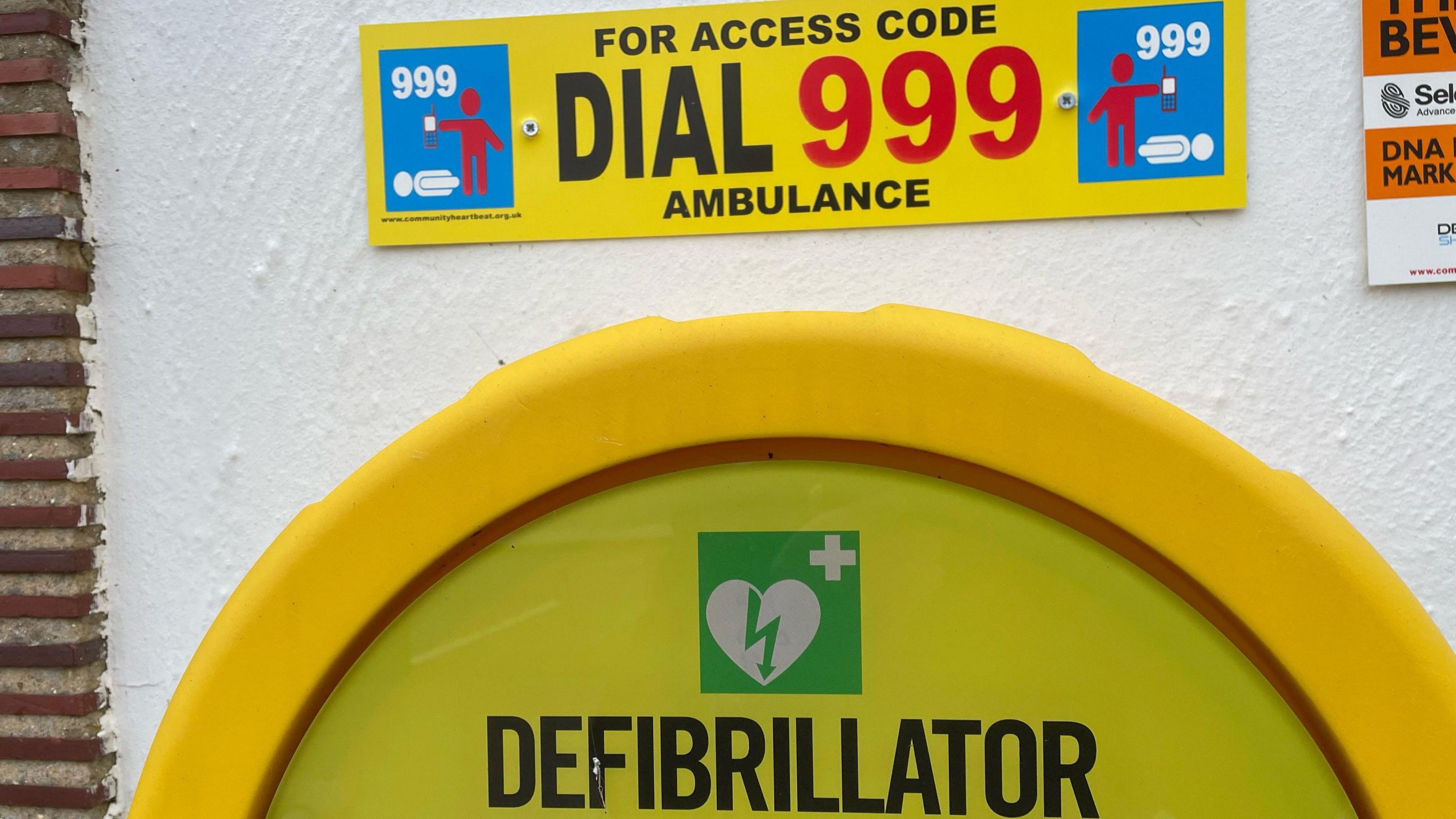 A defibrillator cabinet with a sign saying 'for access code dial 999 ambulance'