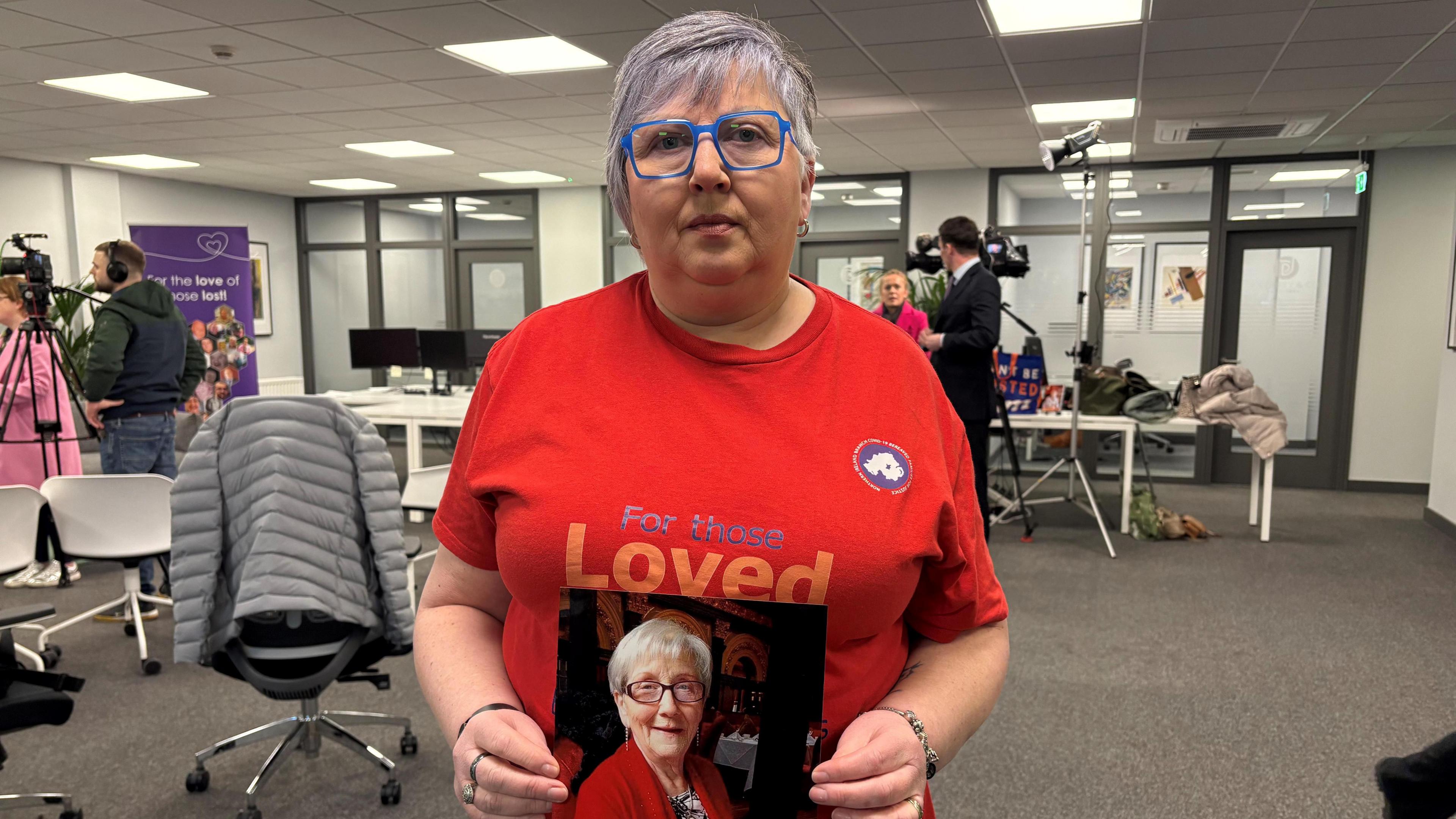 A woman looks at the camera. She is wearing glasses and a red t-shirt, which says 'For those Loved', holds up a photograph of a woman. The woman is in a room with tables and chairs. 