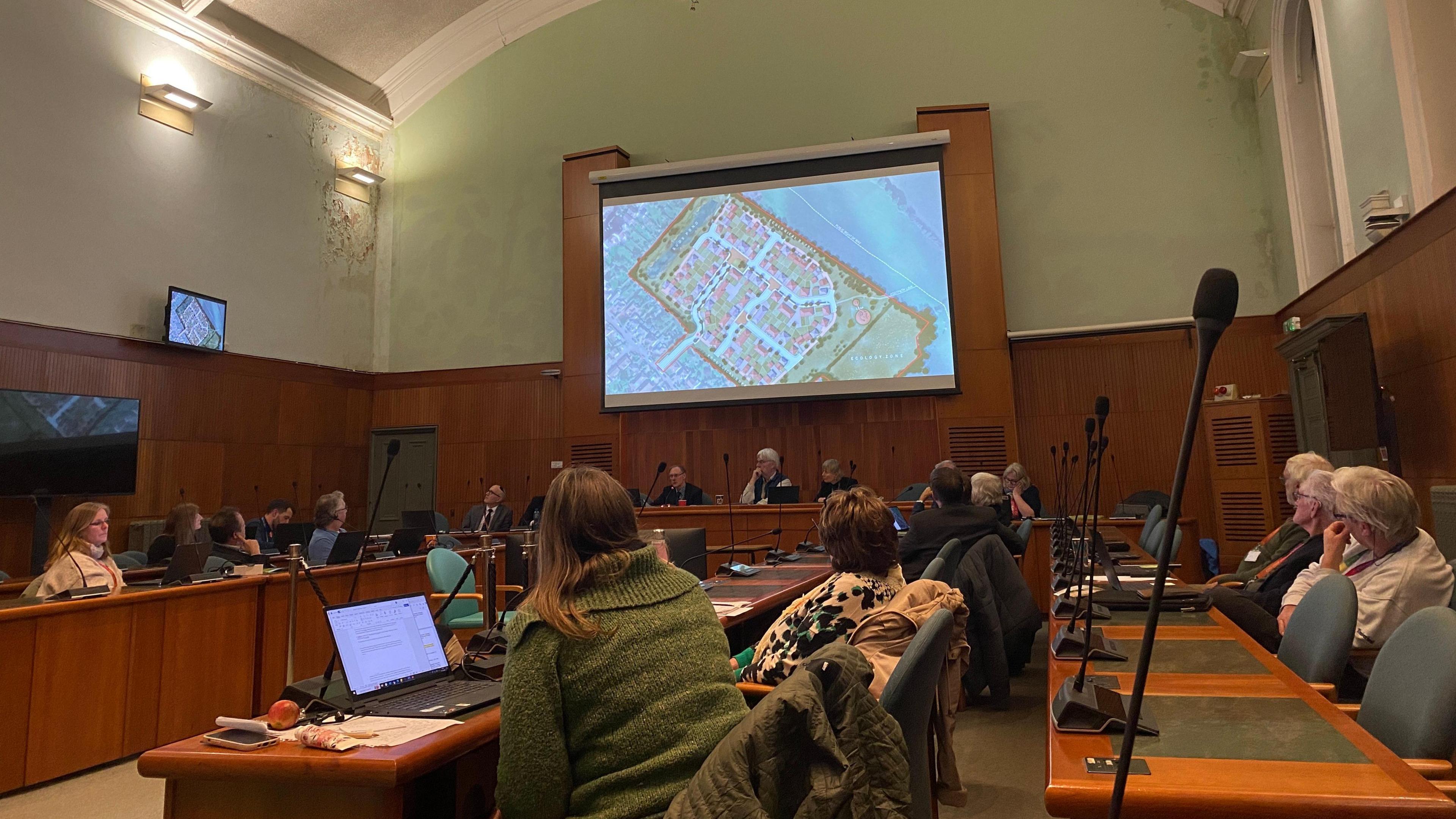 People sit in the North Somerset Council chamber looking up at a large projector screen with a birds-eye plan showing lots of houses on a green space. The room has very high ceilings and is painted a light green, with high wooden panelling running around the outside of the room. 