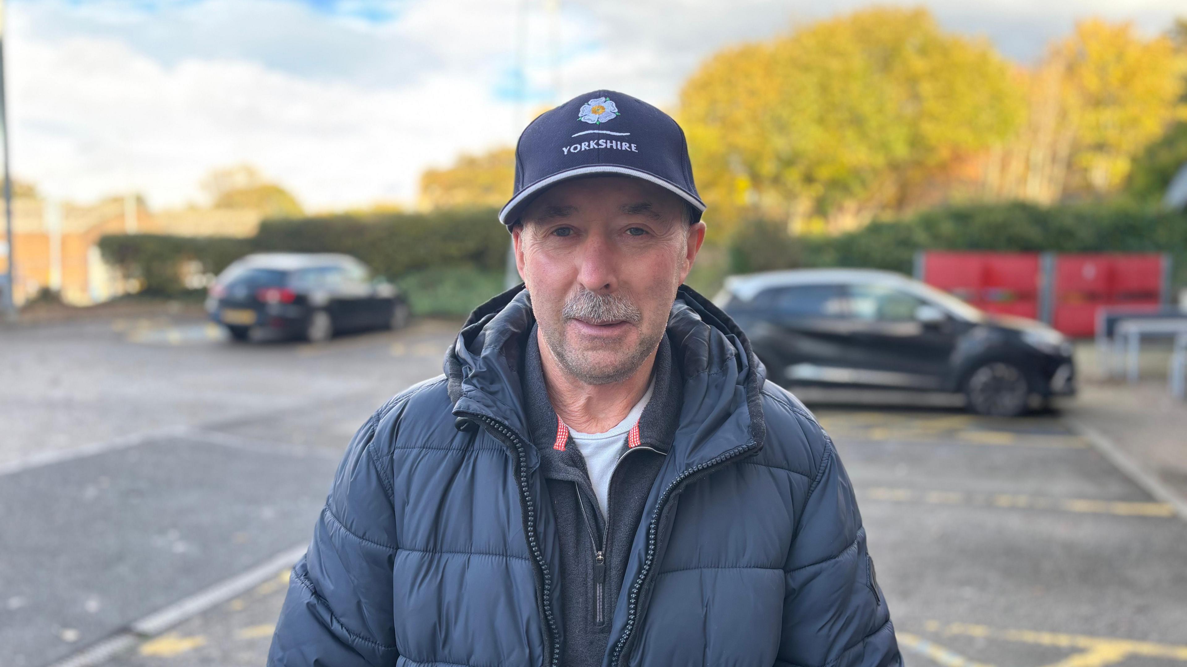 A man in his 60s wearing a navy coat and a baseball cap. He has a small grey moustache and is smiling at the camera.