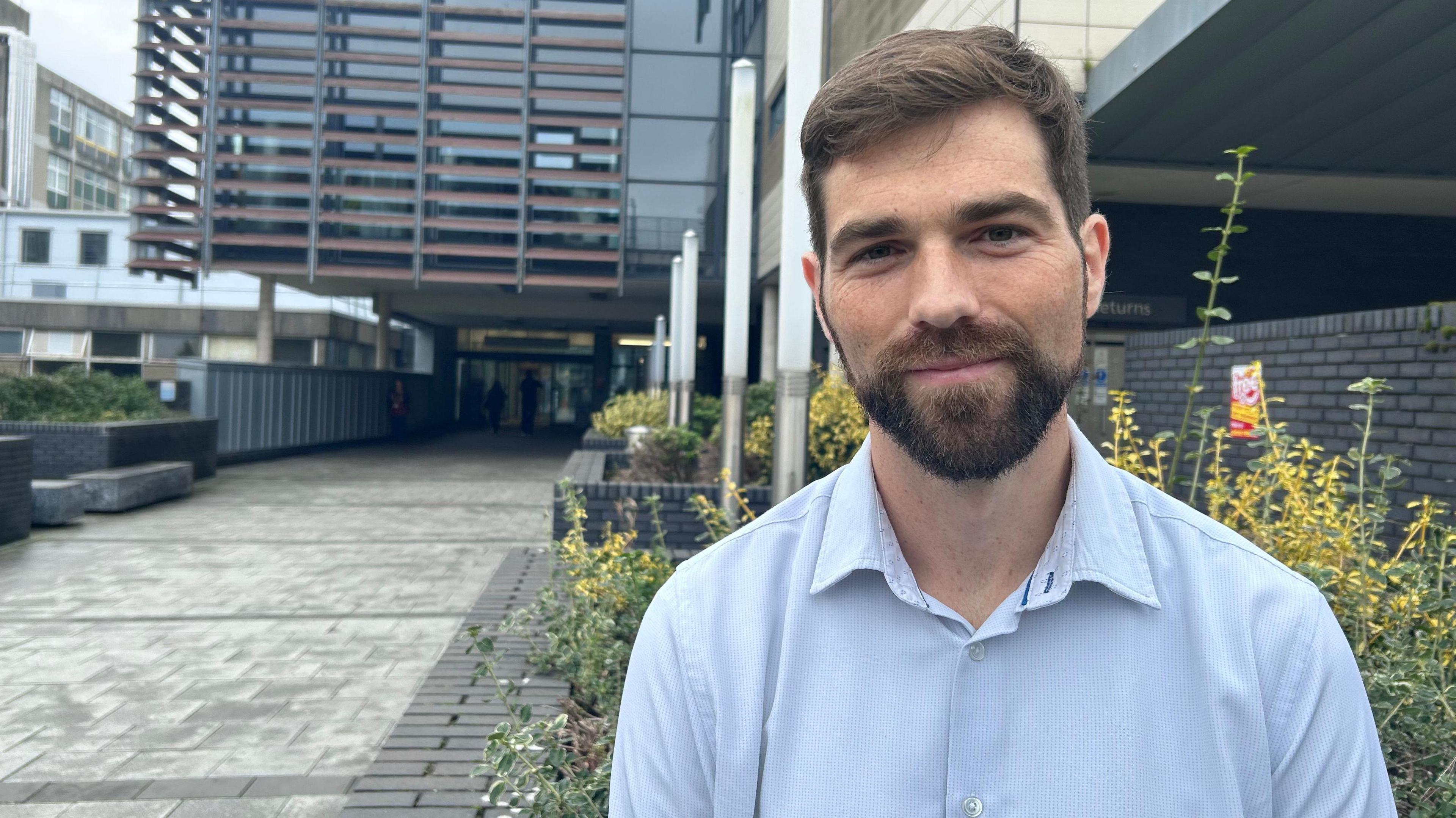 A man is smiling, he has brown hair and a beard. He is wearing a blue shirt.