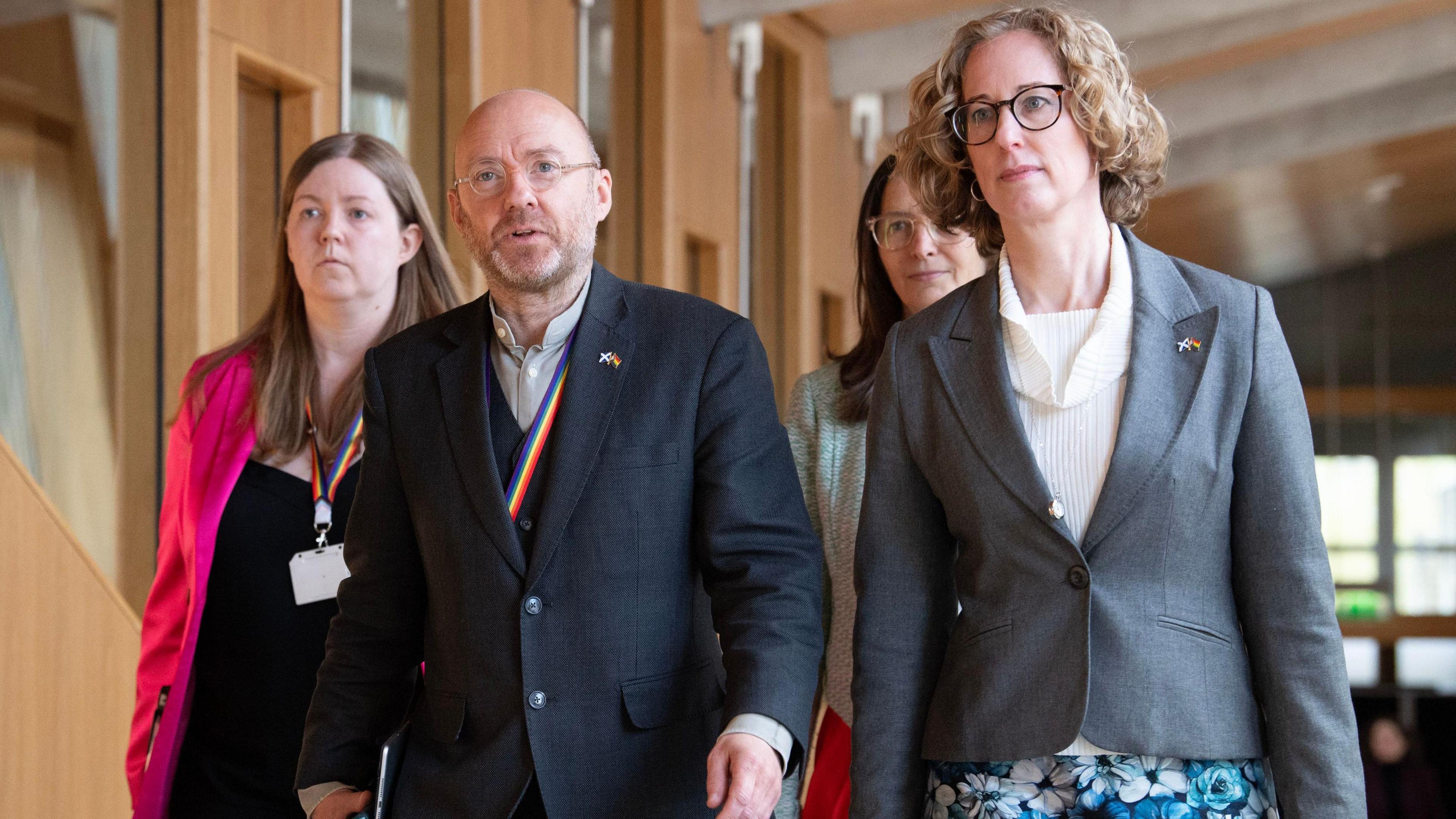 Scottish Green Party co-leaders Patrick Harvie and Lorna Slater photographed walking along a corridor in the Scottish Parliament. Harvie is wearing a dark grey three-piece suit and a lighter coloured shirt. Slater is wearing a white blouse, floral patterned skirt and a grey blazer with a single fastened button.  