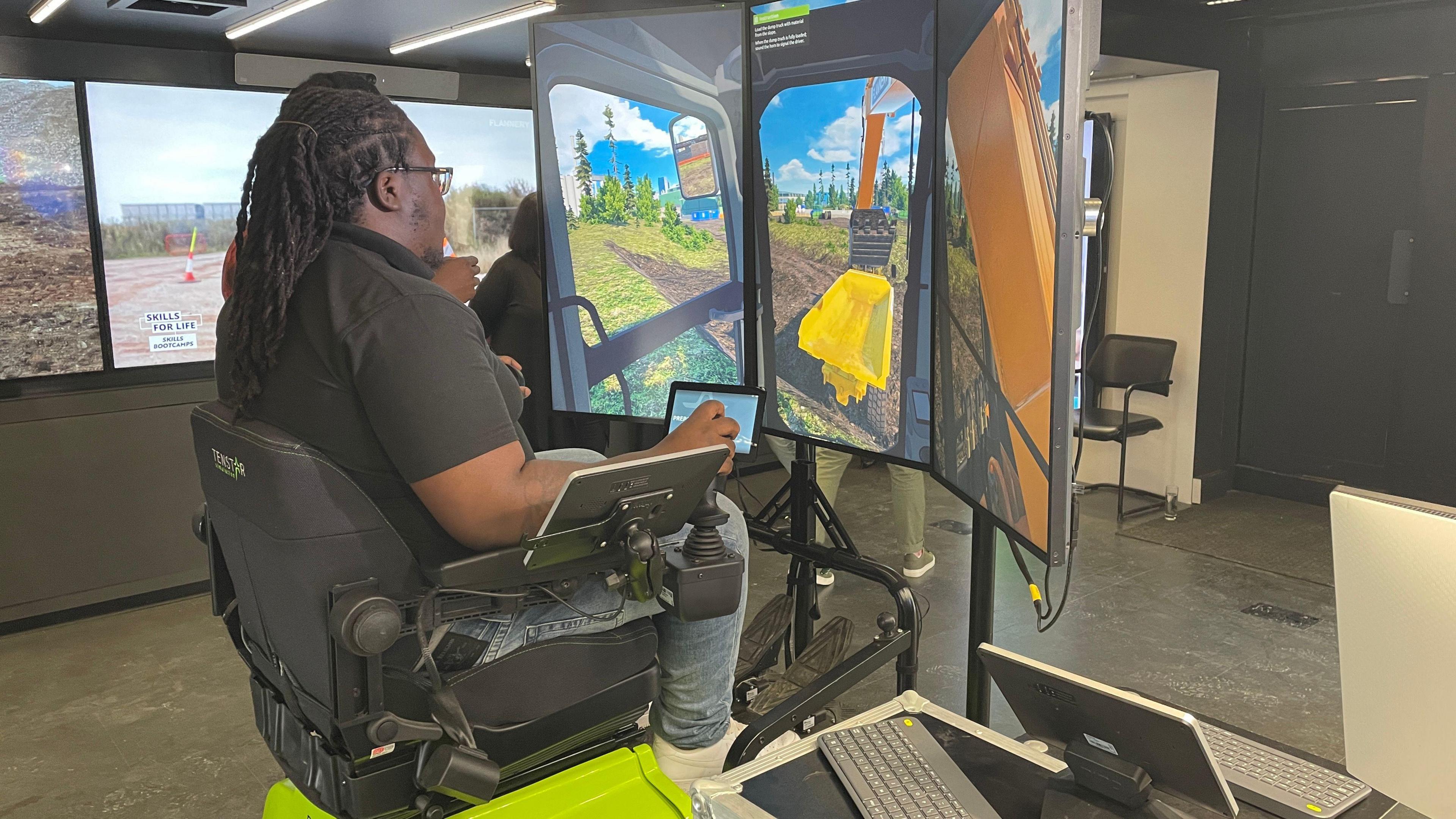 A man sat in a chair in front of three screens which show a simulation as if he is driving a digger