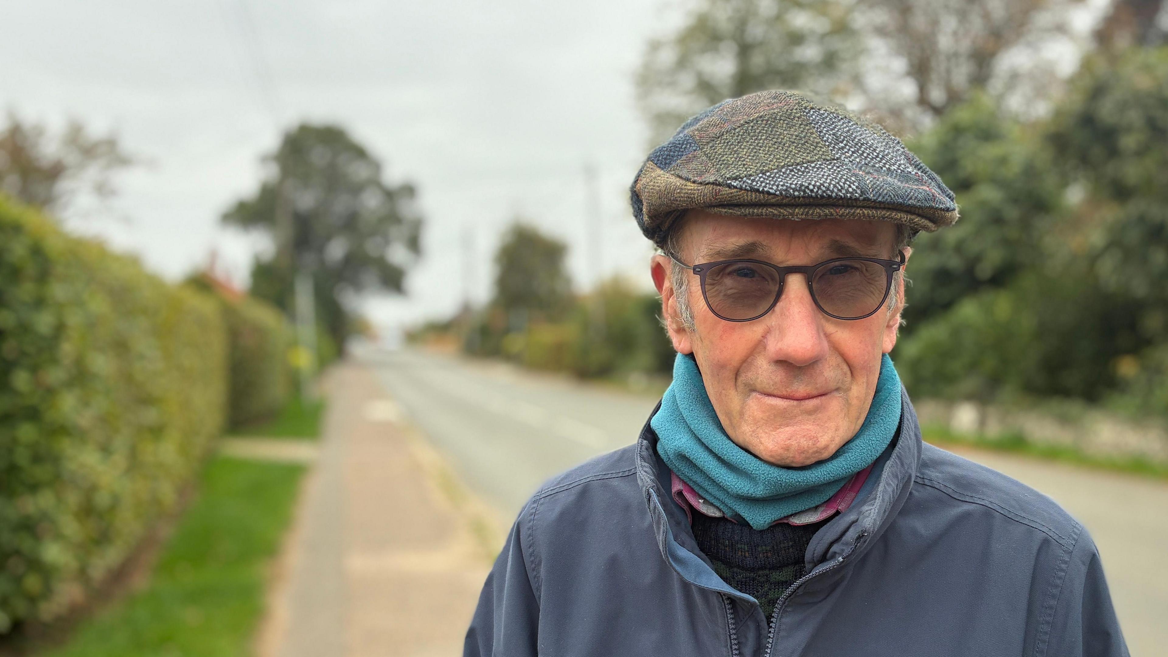 A head and shoulders image of Jim Bootman. He wears a flat cap, turquoise scarf or neckwarmer, a knitted jumper, and a navy jacket on top. There is a narrow residential road behind him which is out of focus.
