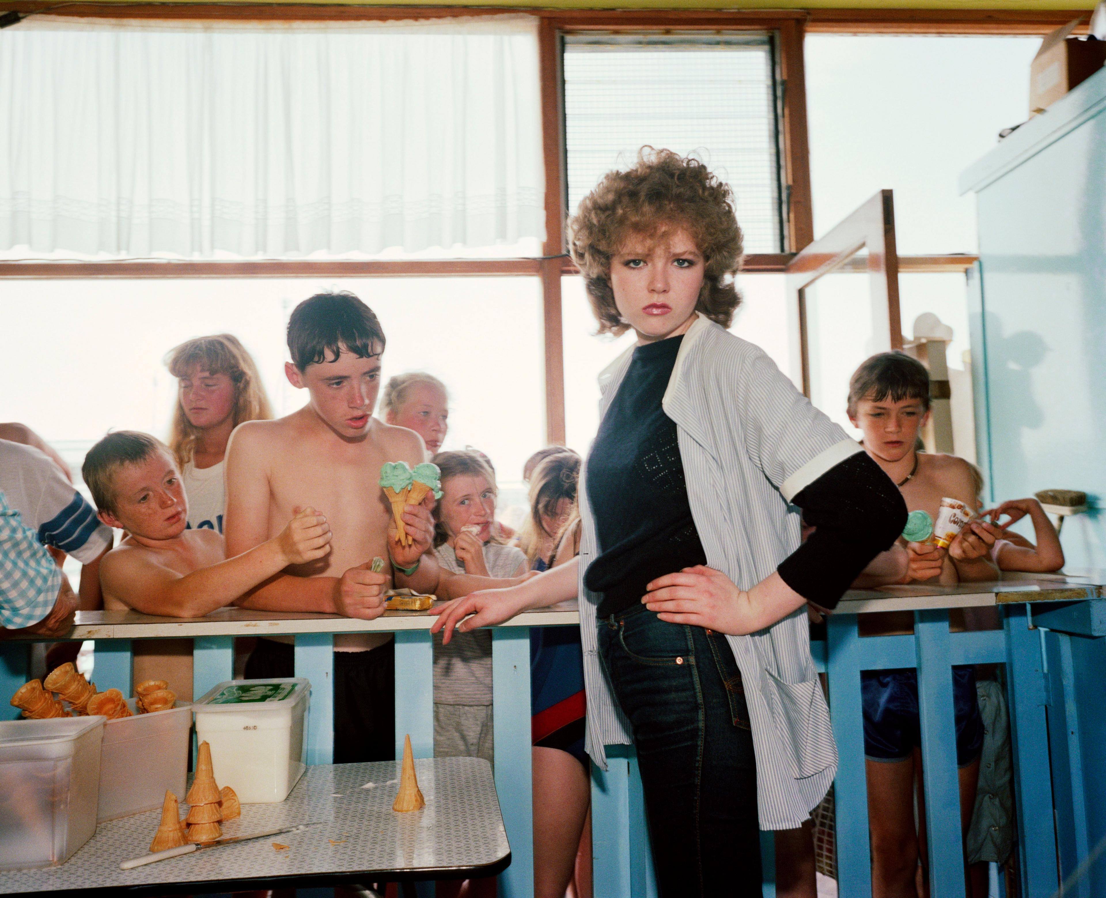 A woman stares at the camera as she takes ice cream orders in a cafe, with kids waiting in the background