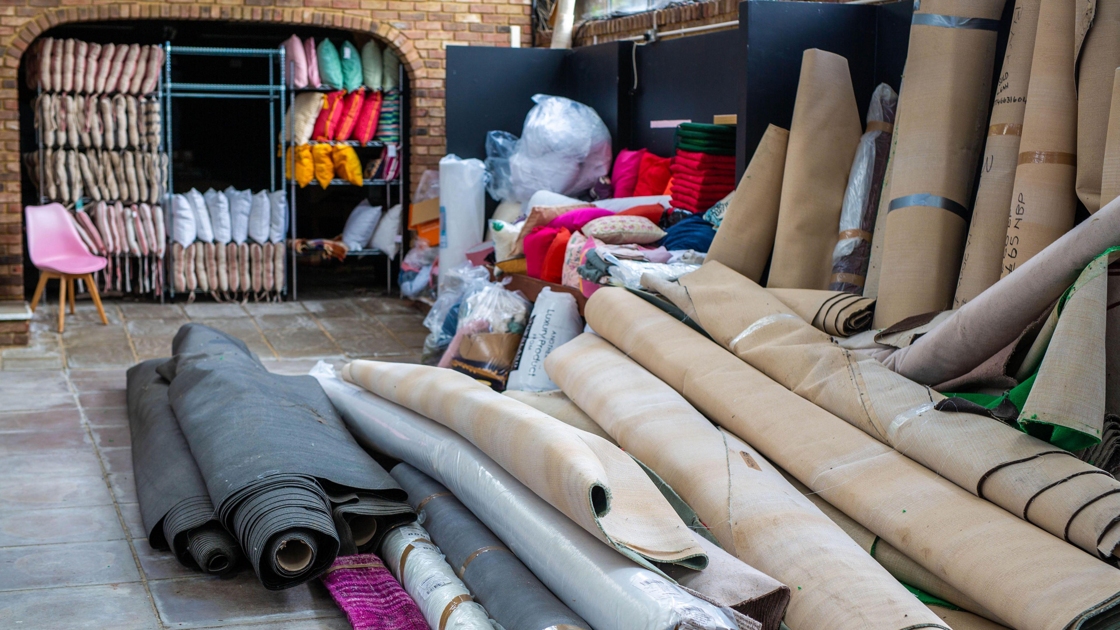 A pile of rolled up carpets on stone tiles. In the background there are shelved with cushions