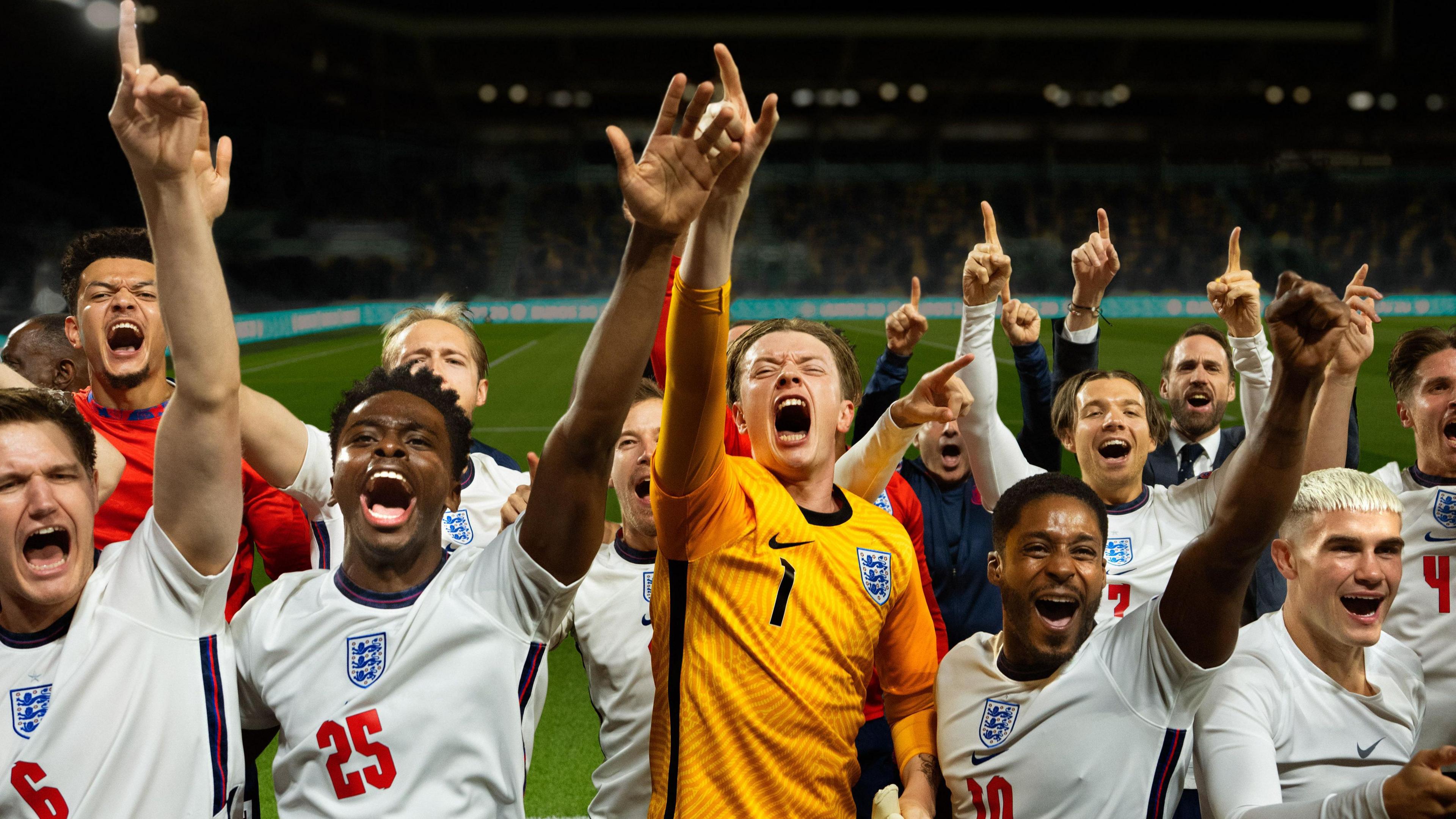 In a scene from Dear England, a team of footballers wearing the England men's team white and red kit celebrate on a football pitch, their goalkeeper, wearing yellow stands in the middle.