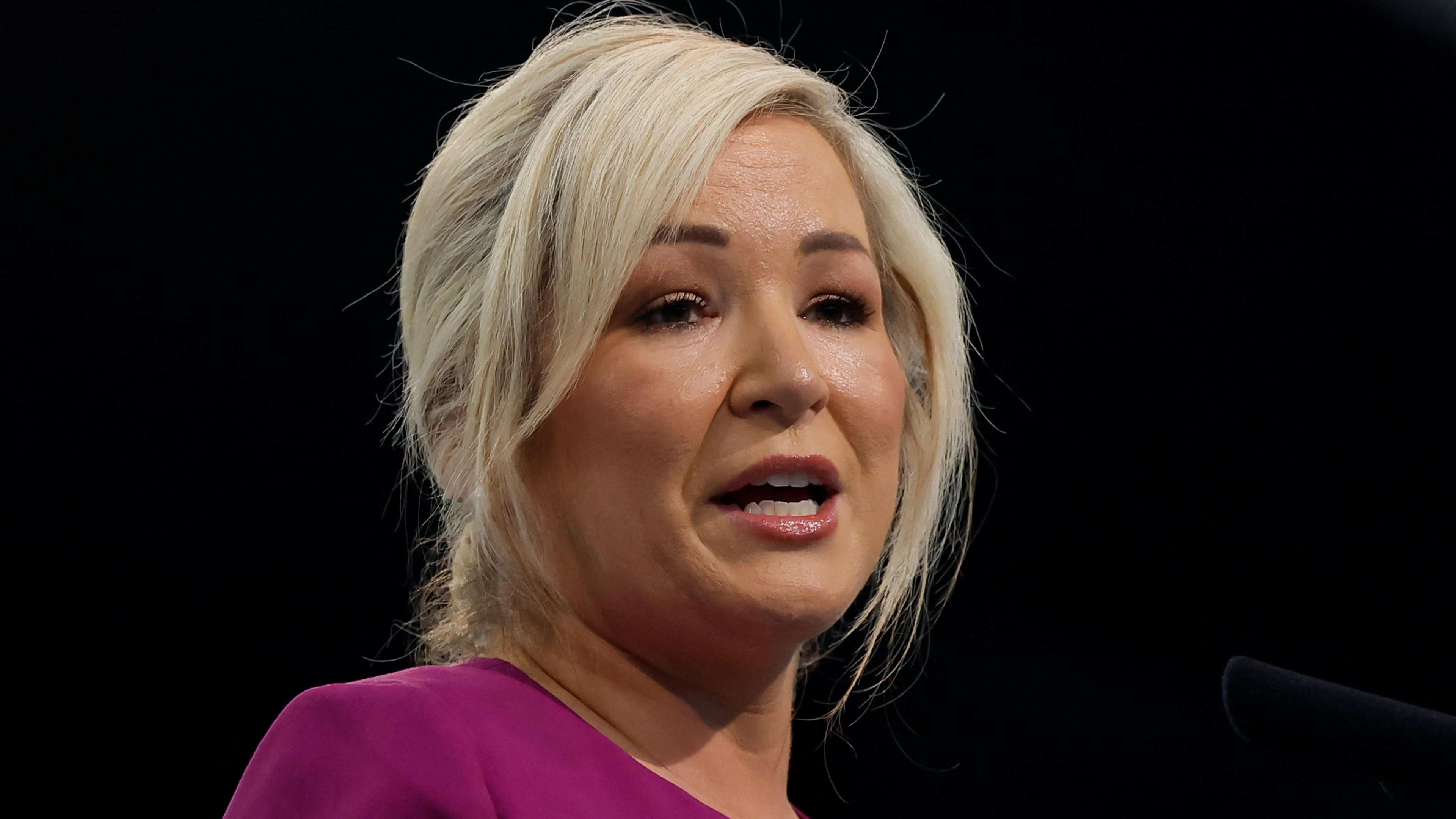 Michelle O'Neill delivers a speech. She's mid-sentence. Her blond hair is tied up and she's wearing a pink jacket over a black dress. The background is black.