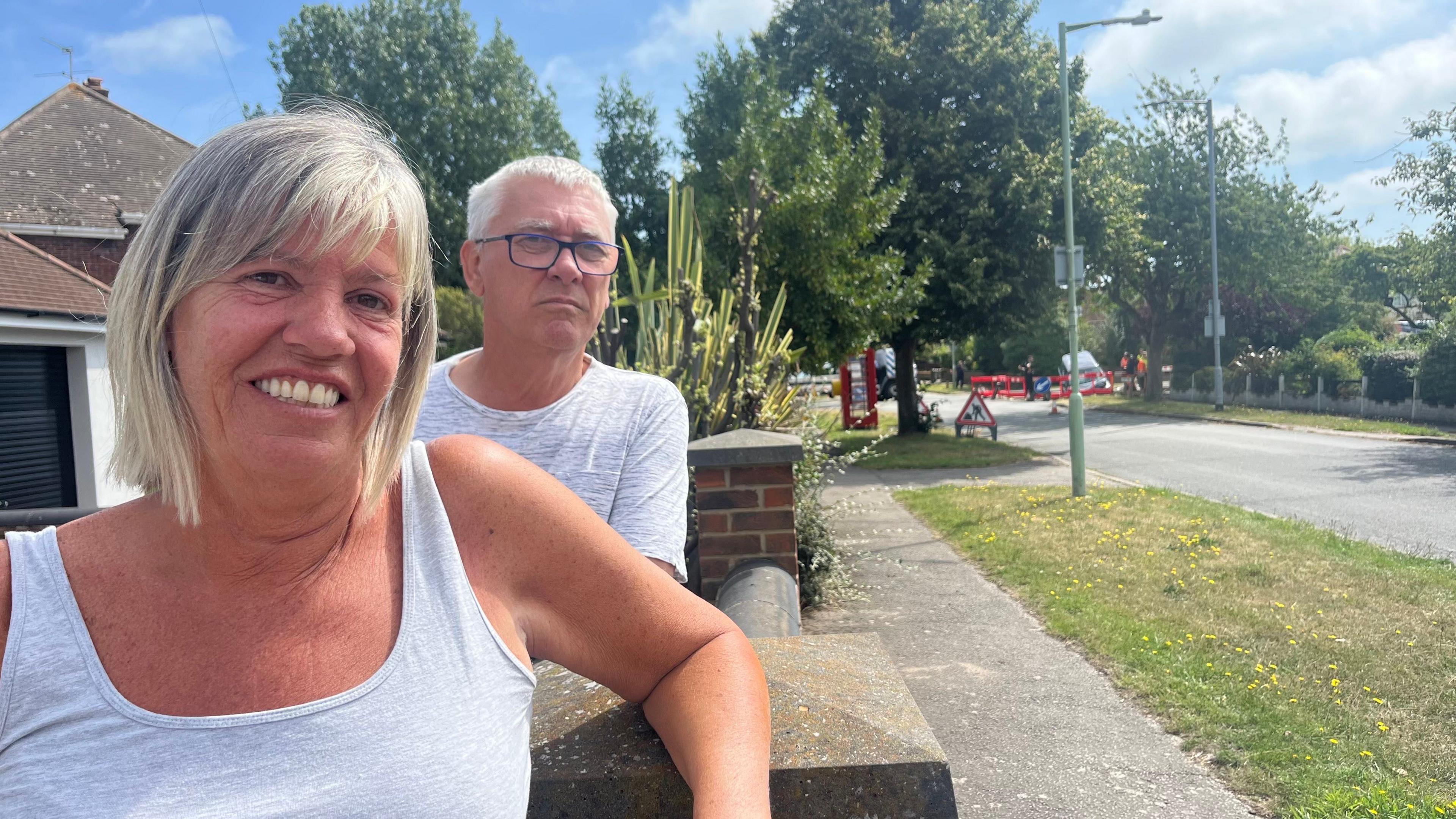 Geoff and Jane May smile at the camera as they rest over their wall in front of their house. Jane has shoulder length grey and blonde hair and wears a grey vest and Geoff has short grey hair and wears a grey T-shirt.