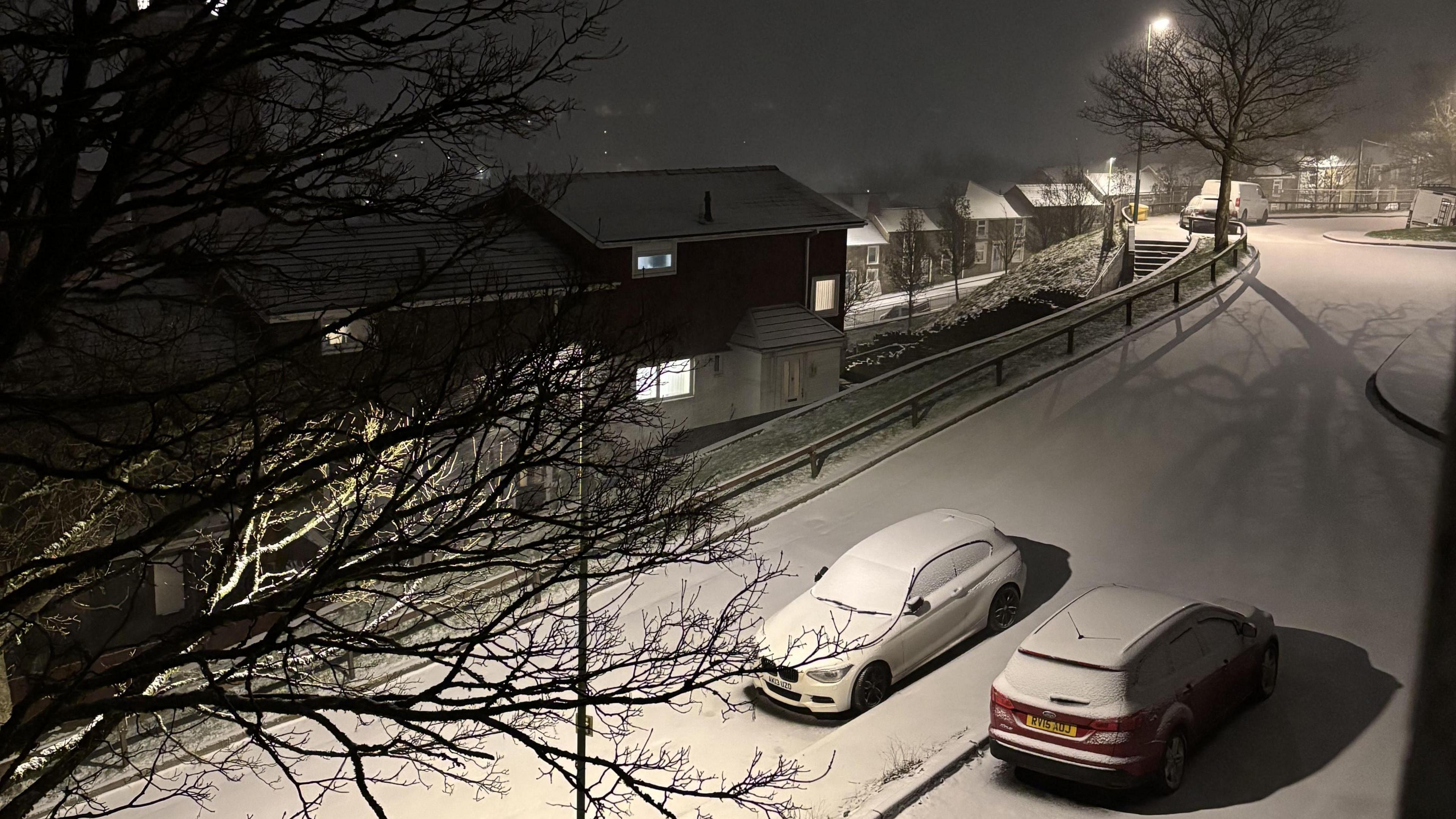 Vehicles are covered in snow in Ebbw Vale as night falls. There is a layer of snow covering the road too meanwhile branches from a tree can be seen on the left side of the image .