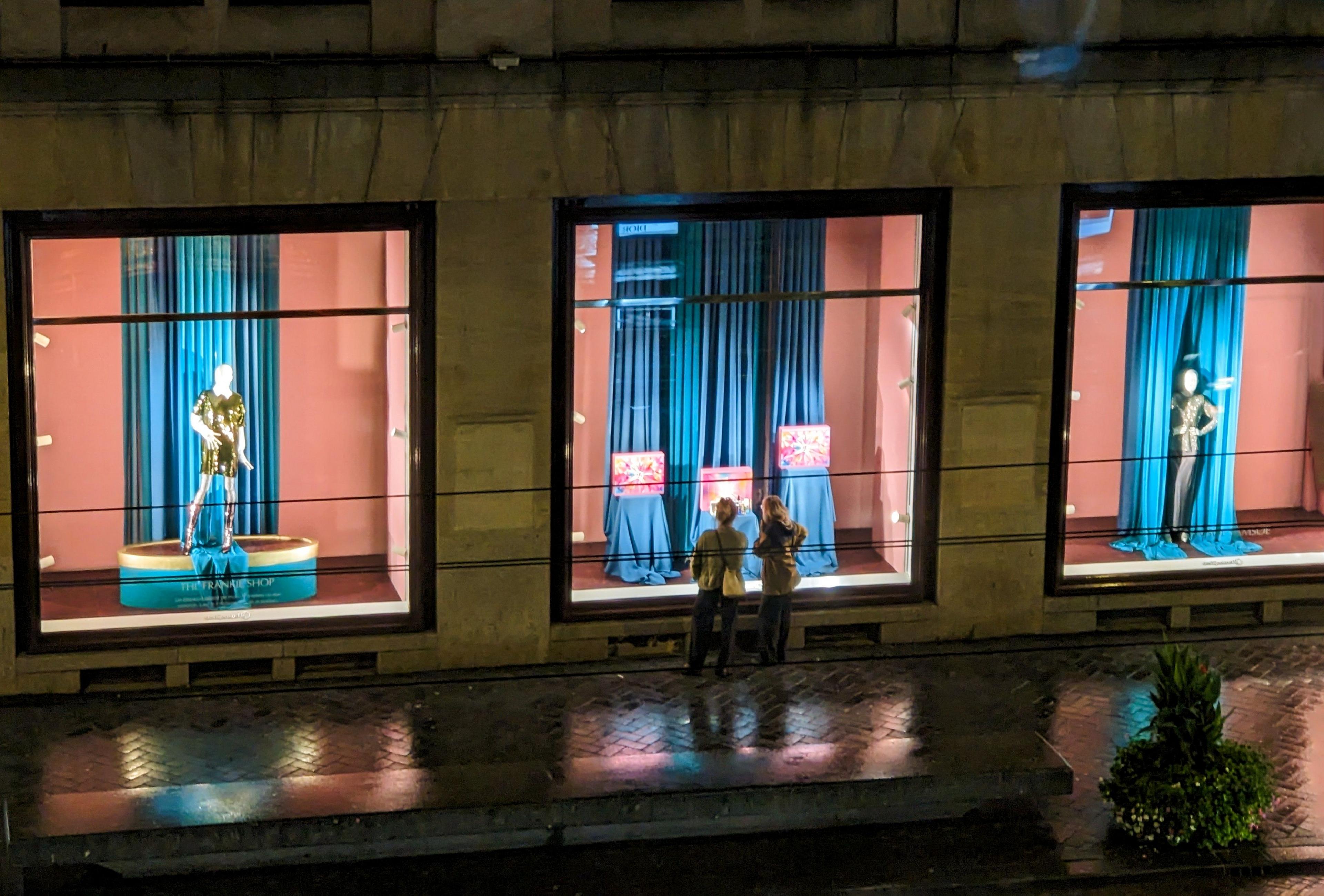 Two people look in the window of a department store at night. There are mannequins in the windows in shiny dresses.